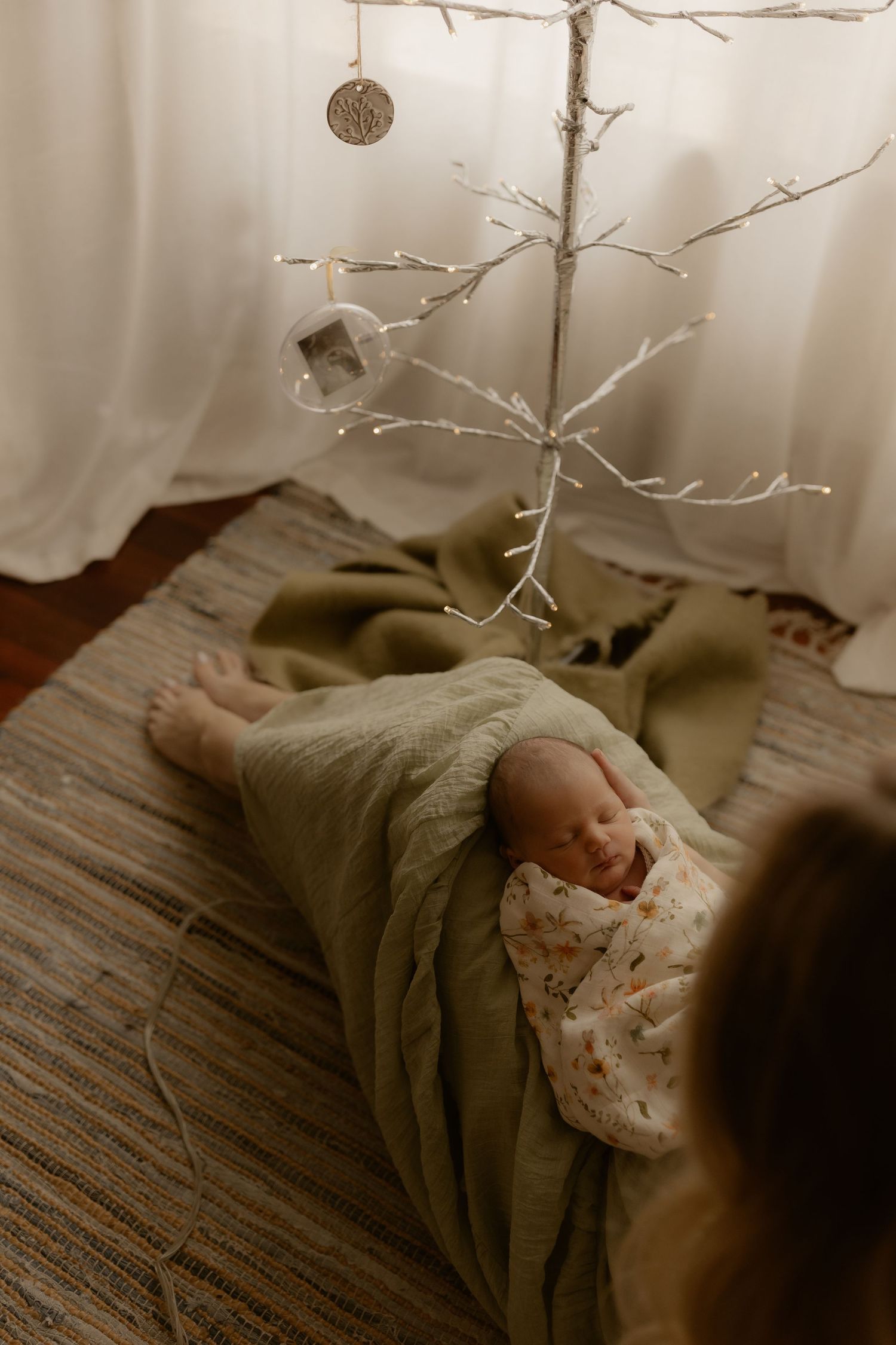 Sleeping newborn on textured brown rug beneath delicate hanging branch decoration.