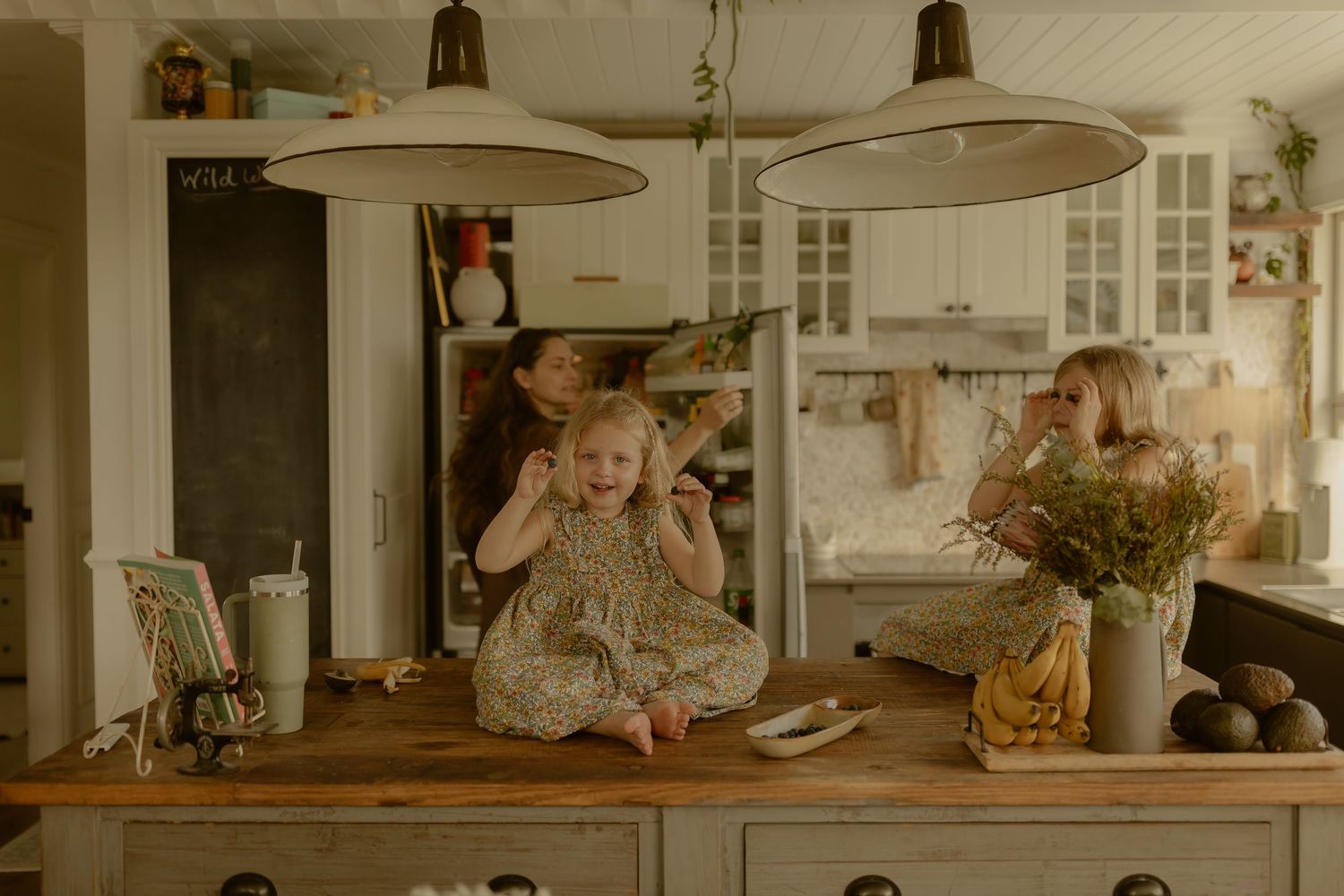 Two young girls play in a charming rustic kitchen with white cabinets and wooden island under pendant lights.