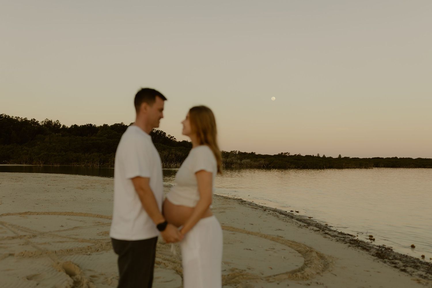 Couple holding hands on a sandy beach at sunset with a calm lake and trees in the background.
