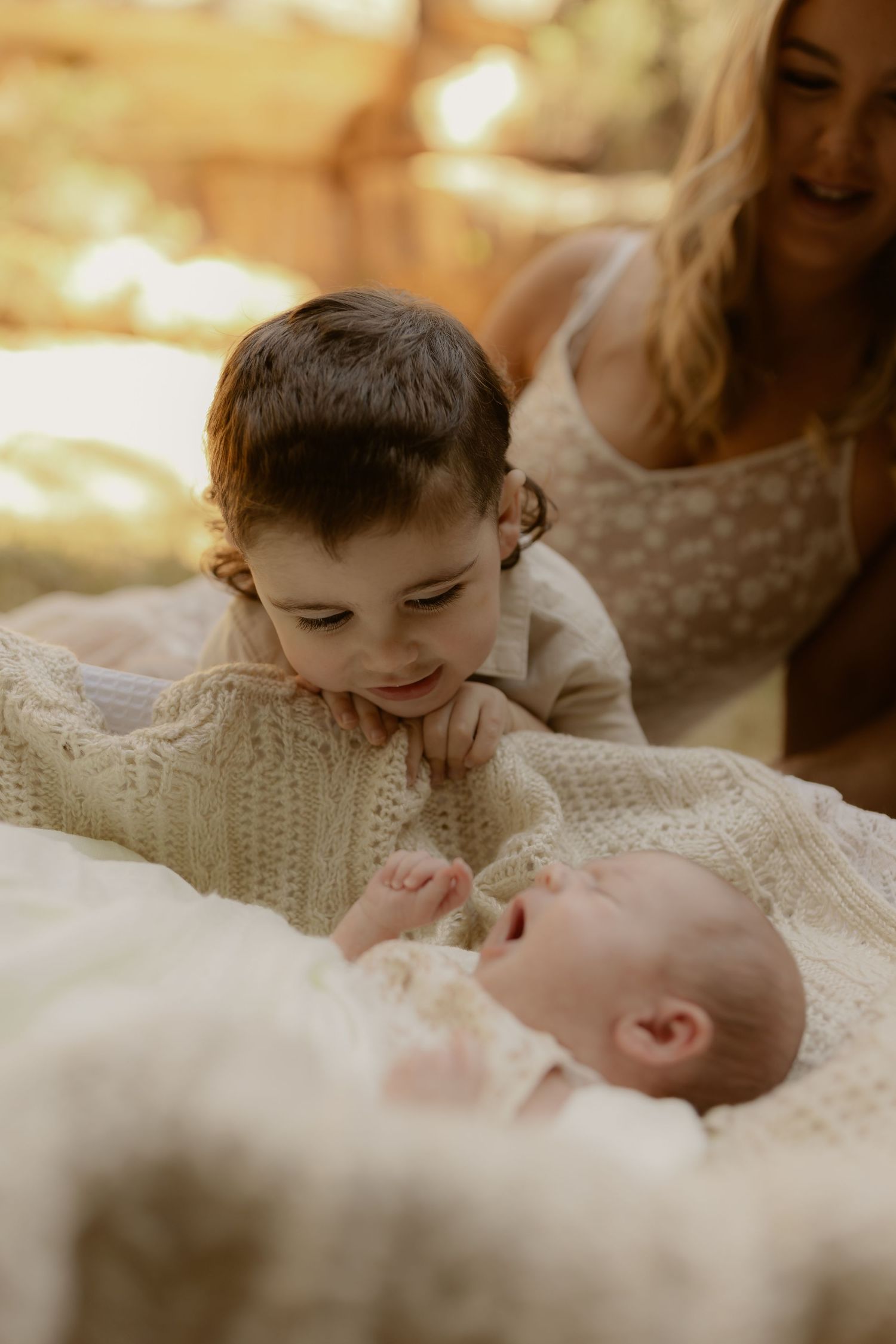 Young child gazes lovingly at newborn baby while mother smiles warmly in golden light.