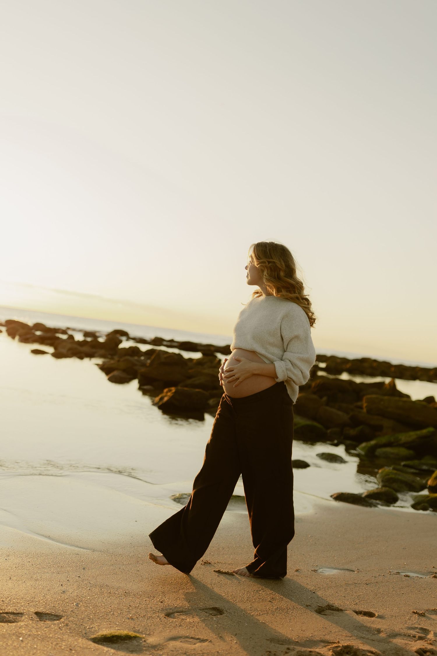 Pregnant woman in white sweater holding belly while walking barefoot on beach at sunset.