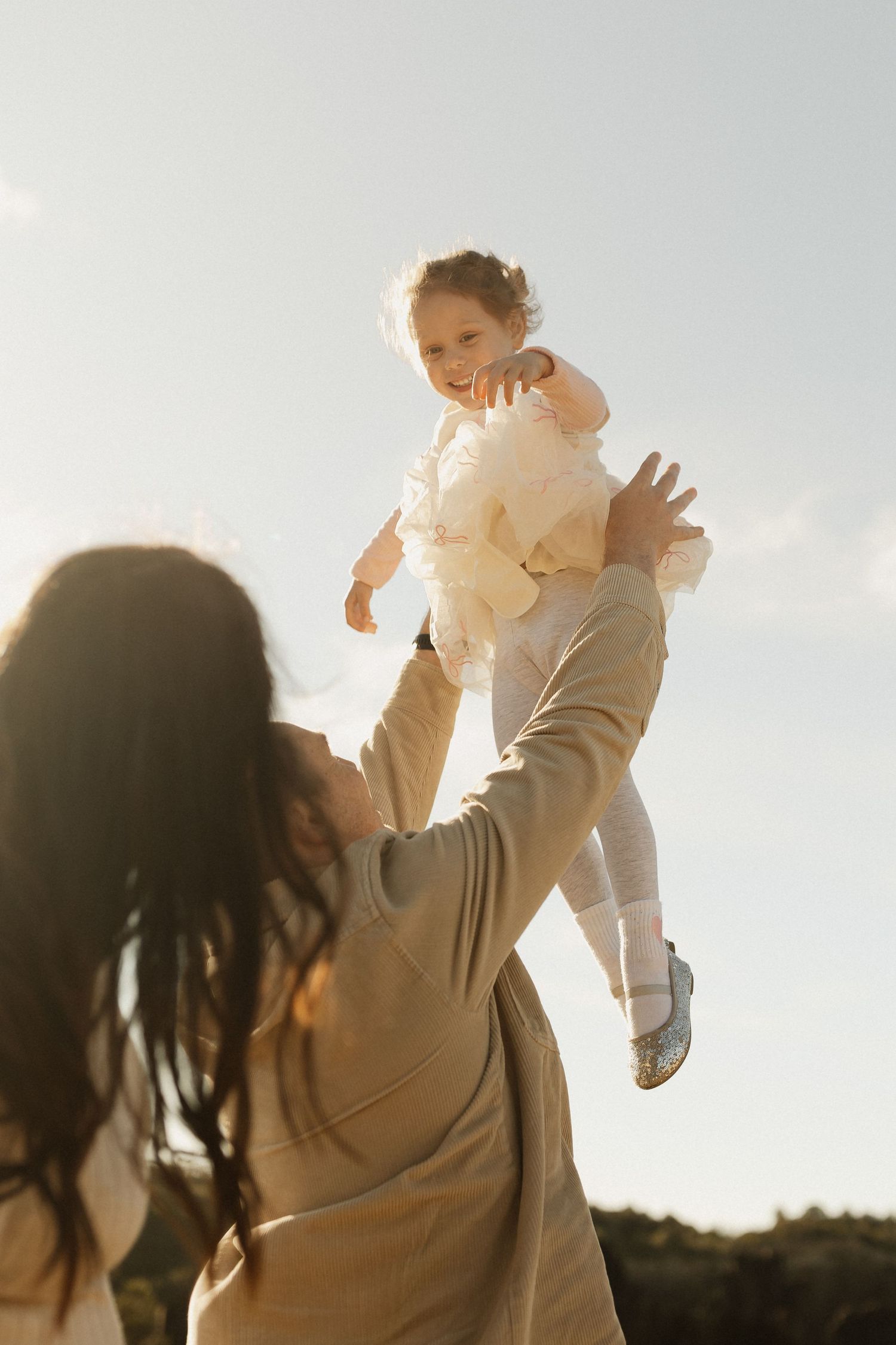 Playful moment as parent lifts child high into the air against bright sky.