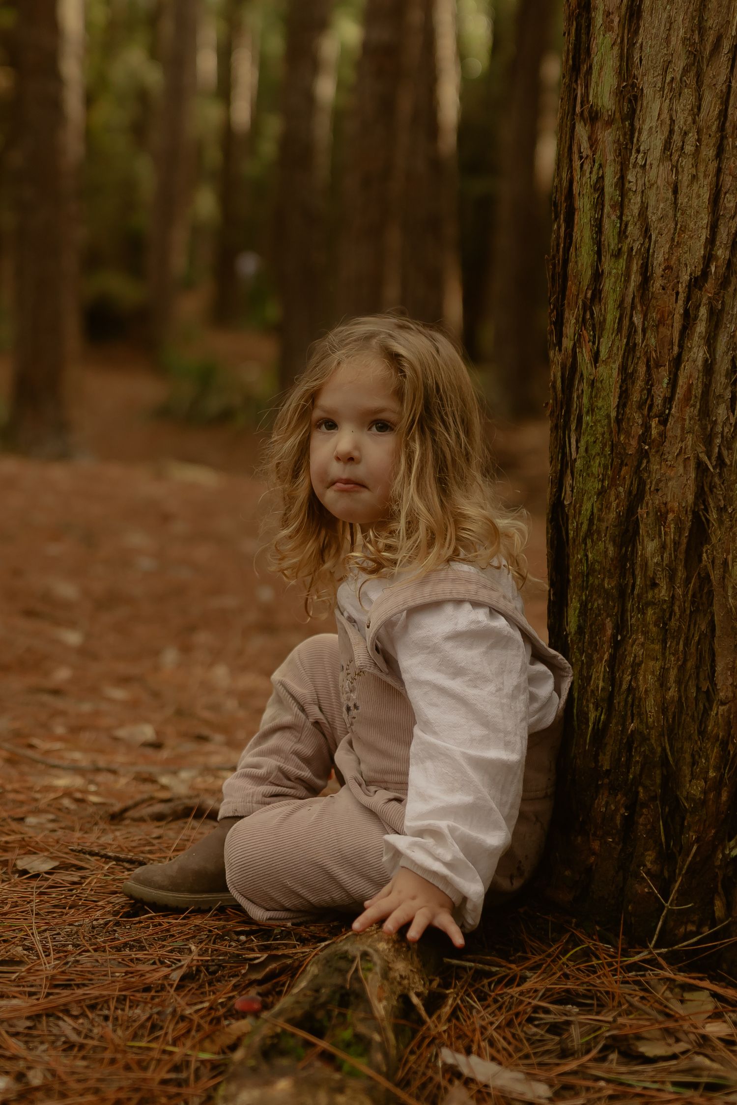 A young curly-haired girl in neutral tones sits against a pine tree on a forest floor covered in fallen needles.