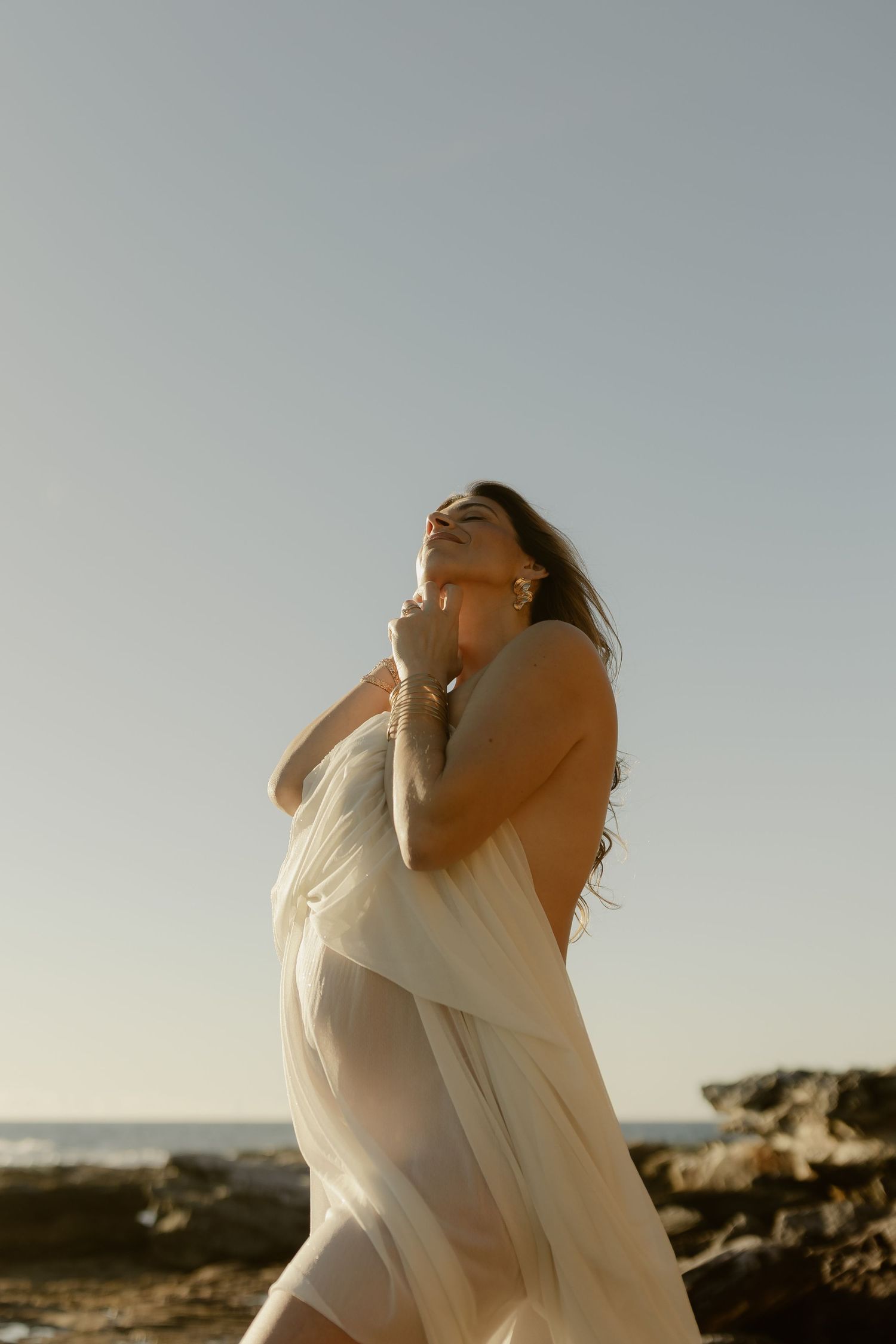 Woman in sheer white dress poses on rocky beach at sunset, hair flowing, looking up with hands clasped.