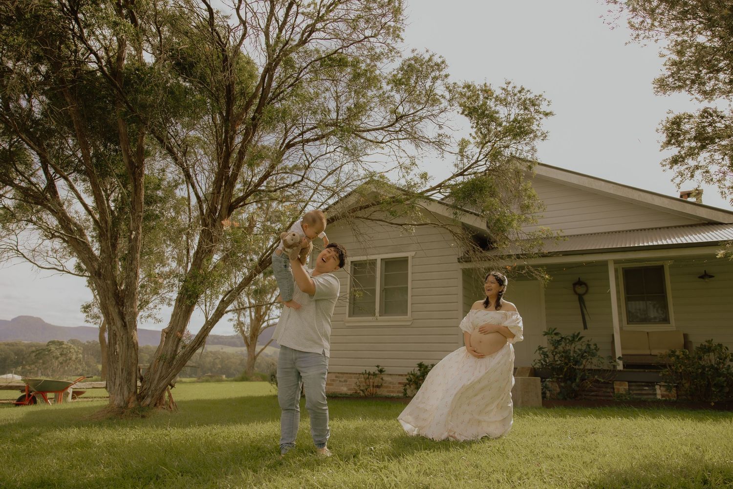 Couple dancing joyfully on green lawn in front of charming white farmhouse surrounded by trees.