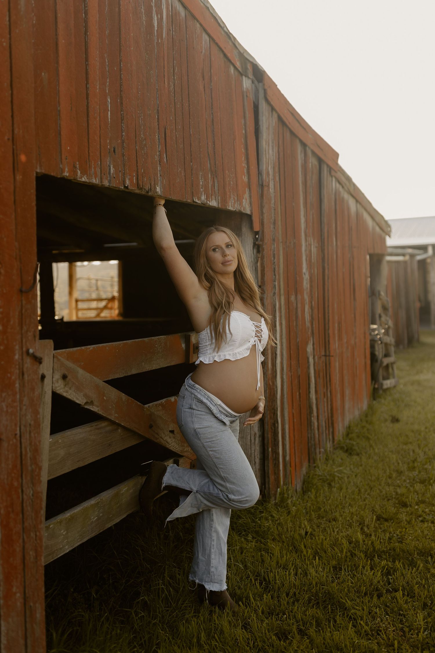 Pregnant woman in white crop top and jeans leaning against rustic red barn door on a farm at golden hour.
