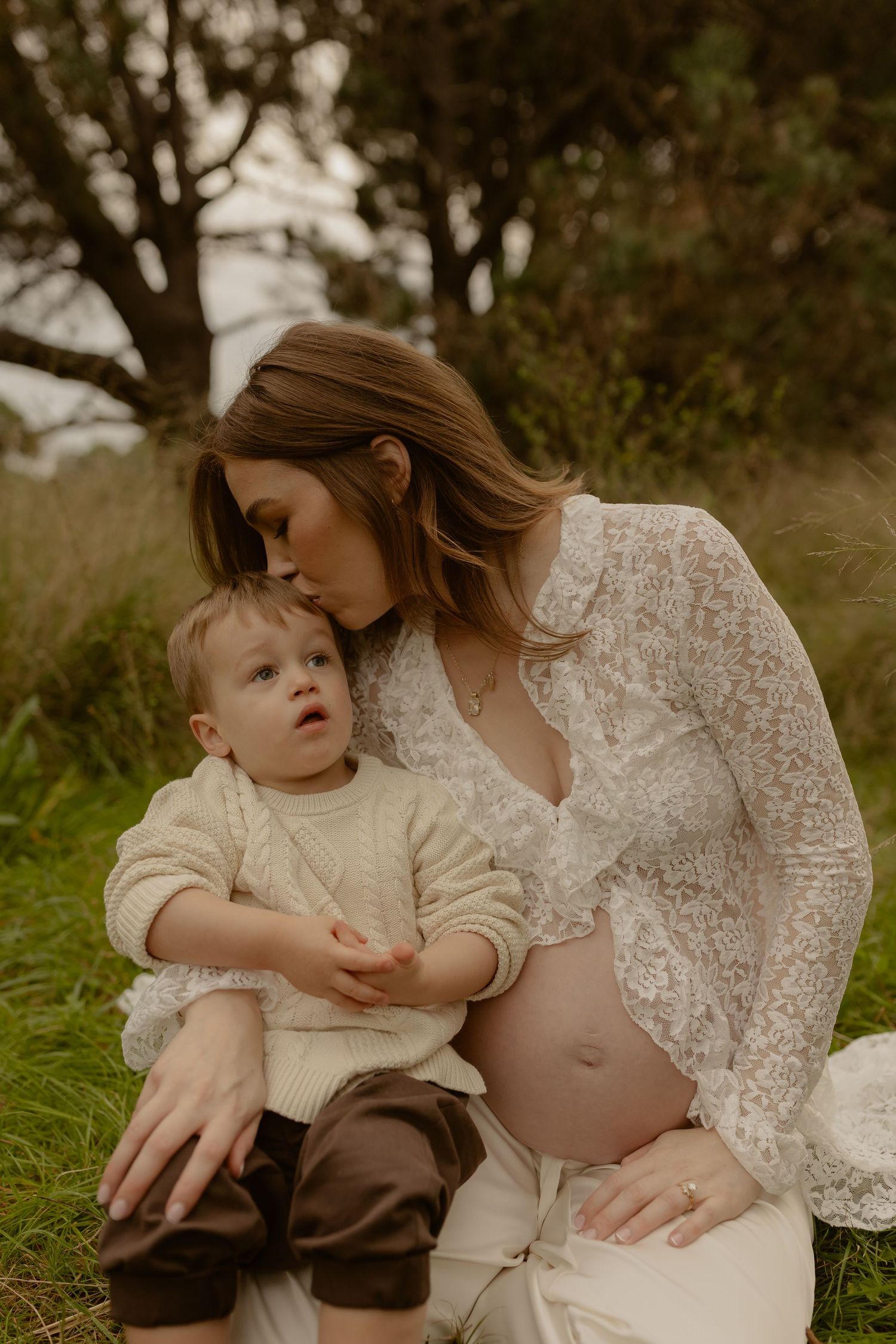 A pregnant woman in a white lace dress tenderly kisses her toddler son while sitting on grass outdoors.
