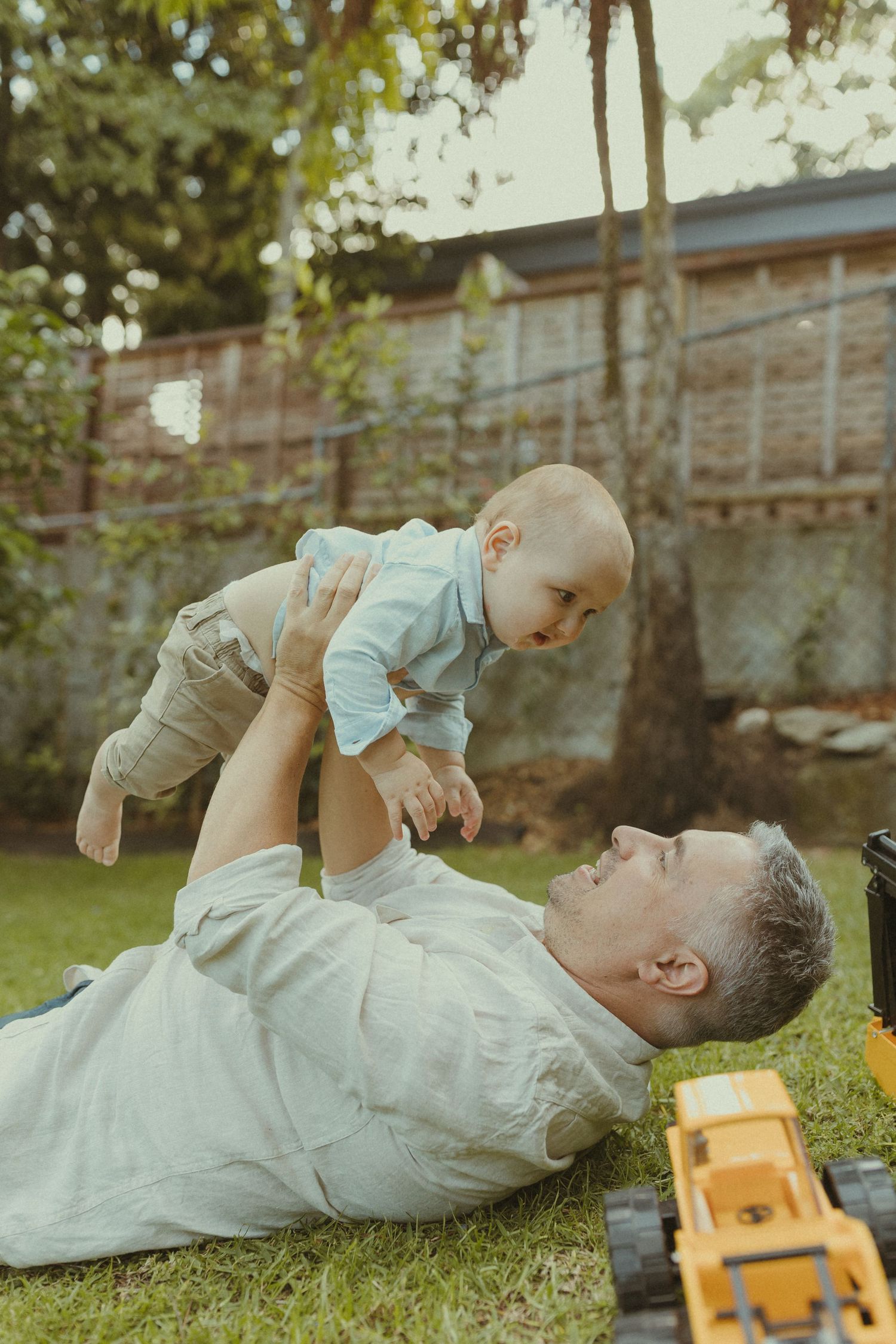 A tender moment between parent and baby lying on grass in a backyard garden setting.