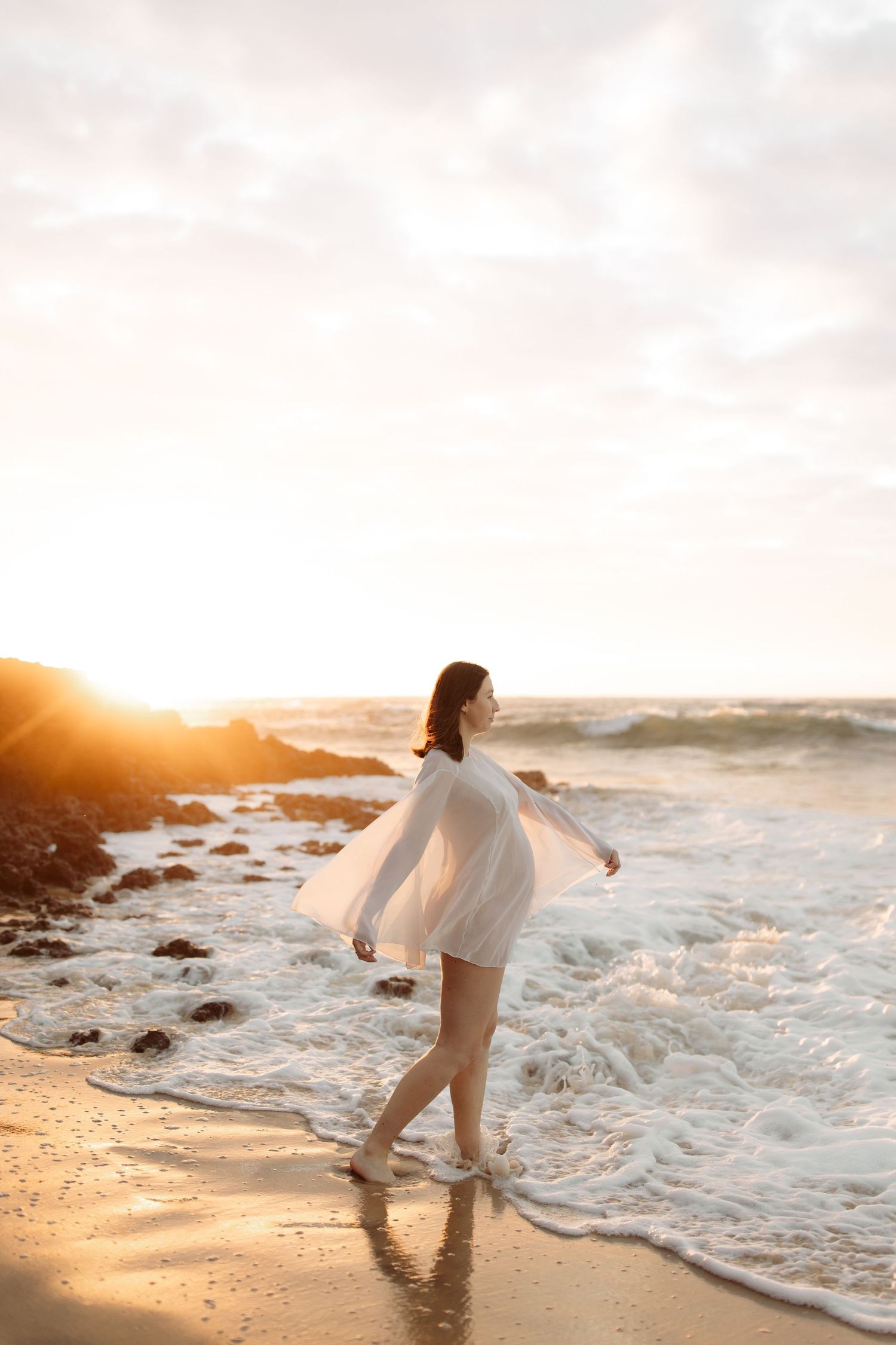 Person in white dress dances along the beach shoreline during golden sunset as waves crash nearby.