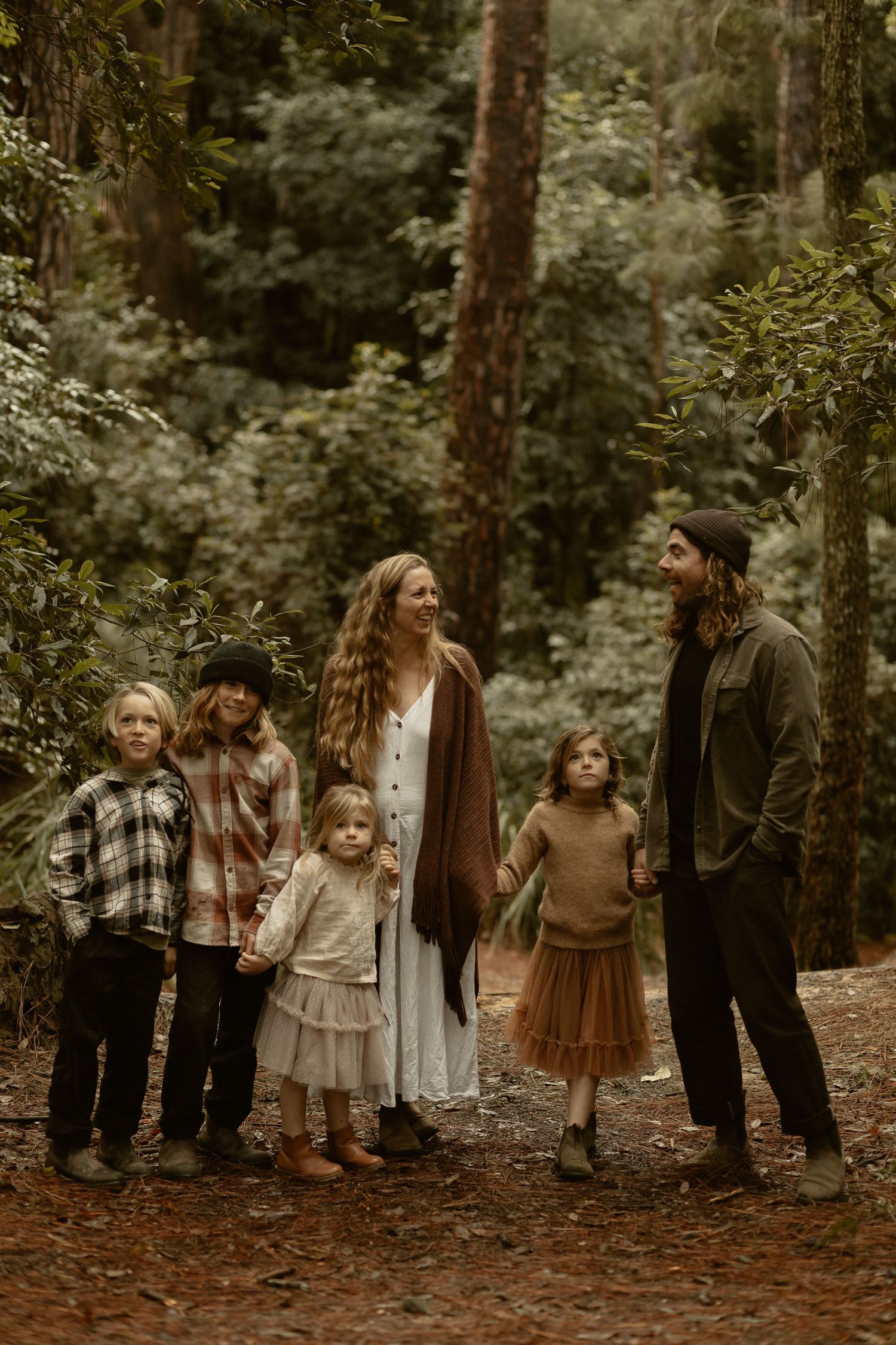 Group wearing vintage-inspired clothing poses together in a moody forest setting with muted earth tones.