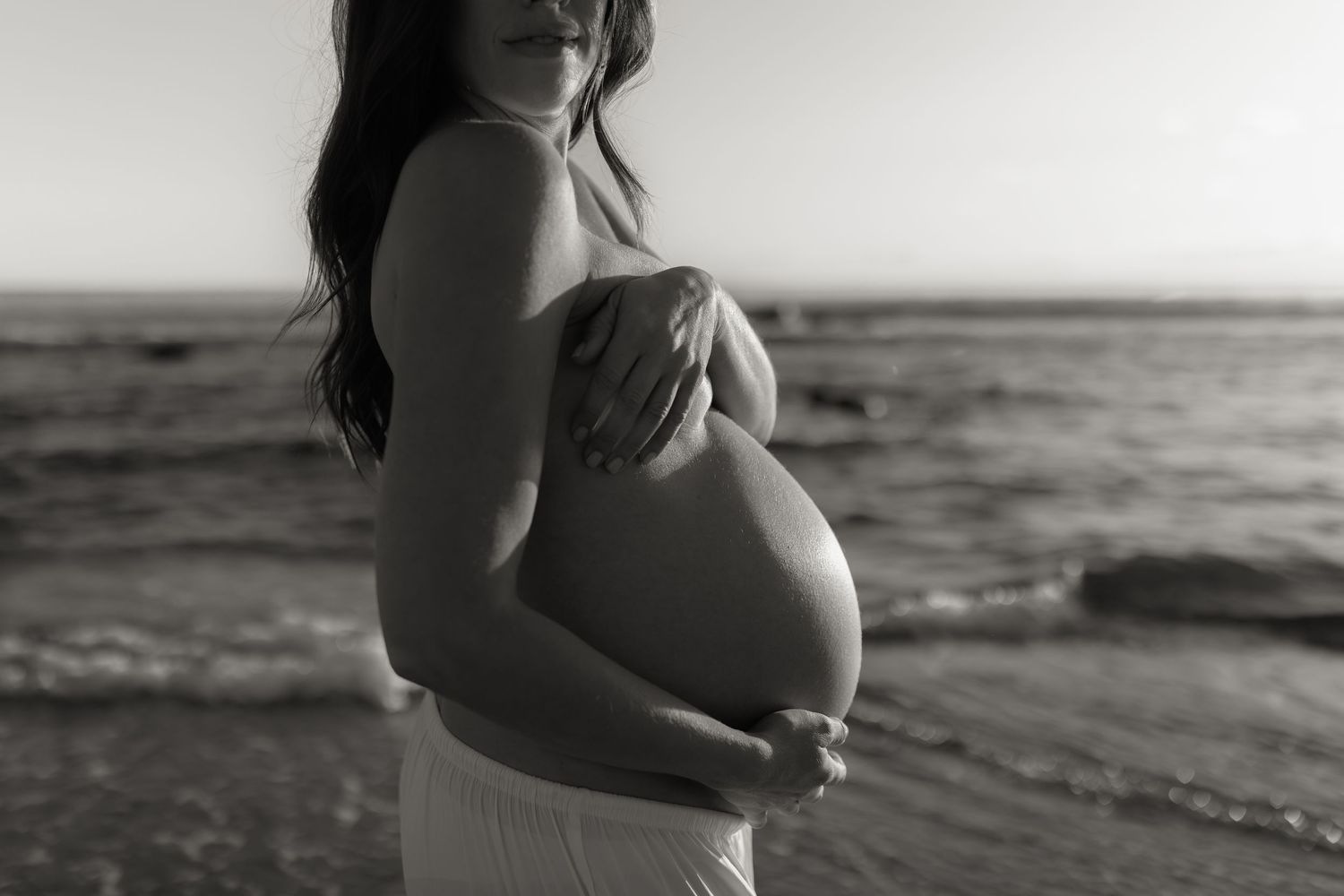 Black and white photo of expecting couple sharing intimate moment on beach at dusk.