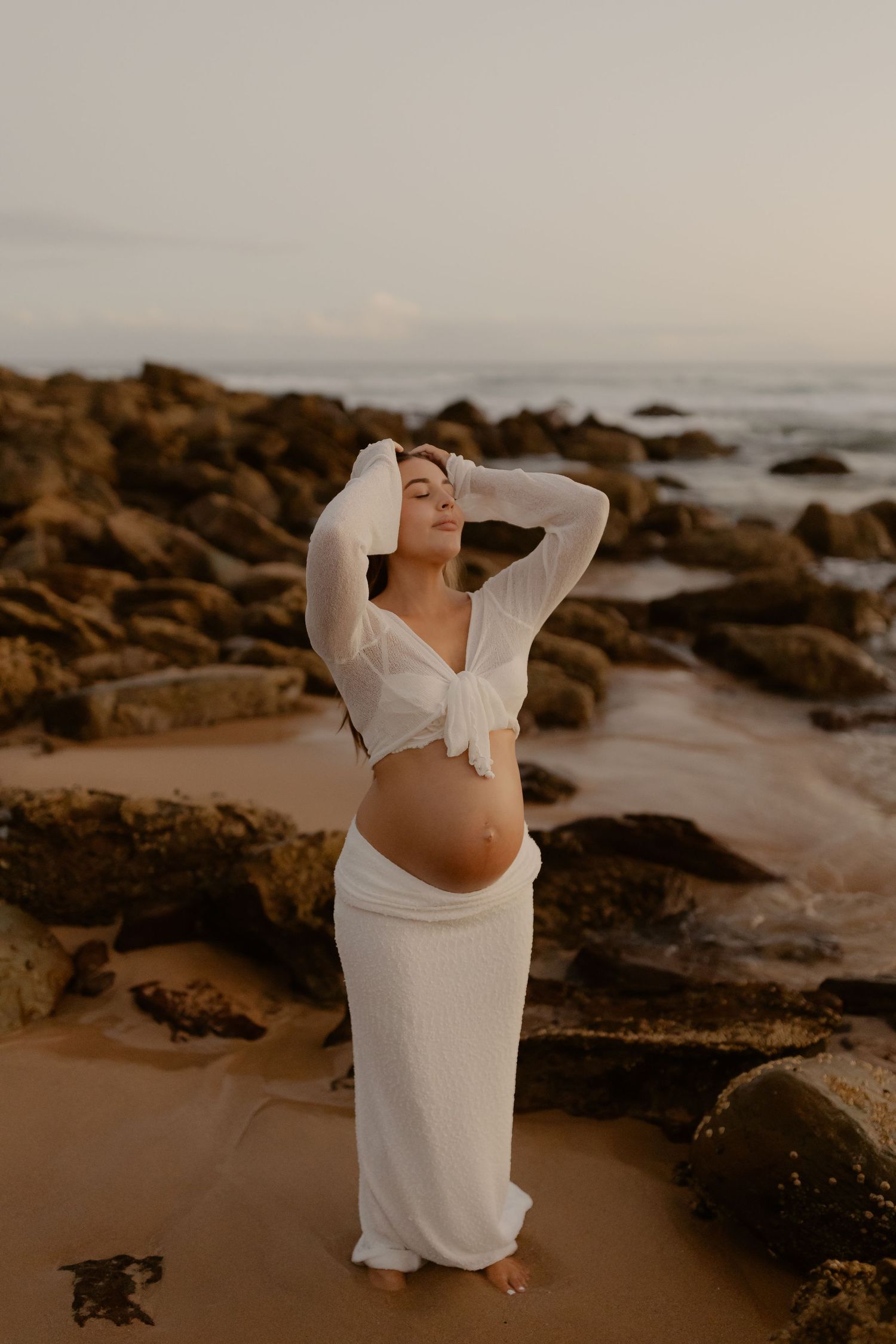 A person in a flowing white dress poses gracefully on a rocky beach at sunset creating a dreamy coastal atmosphere.