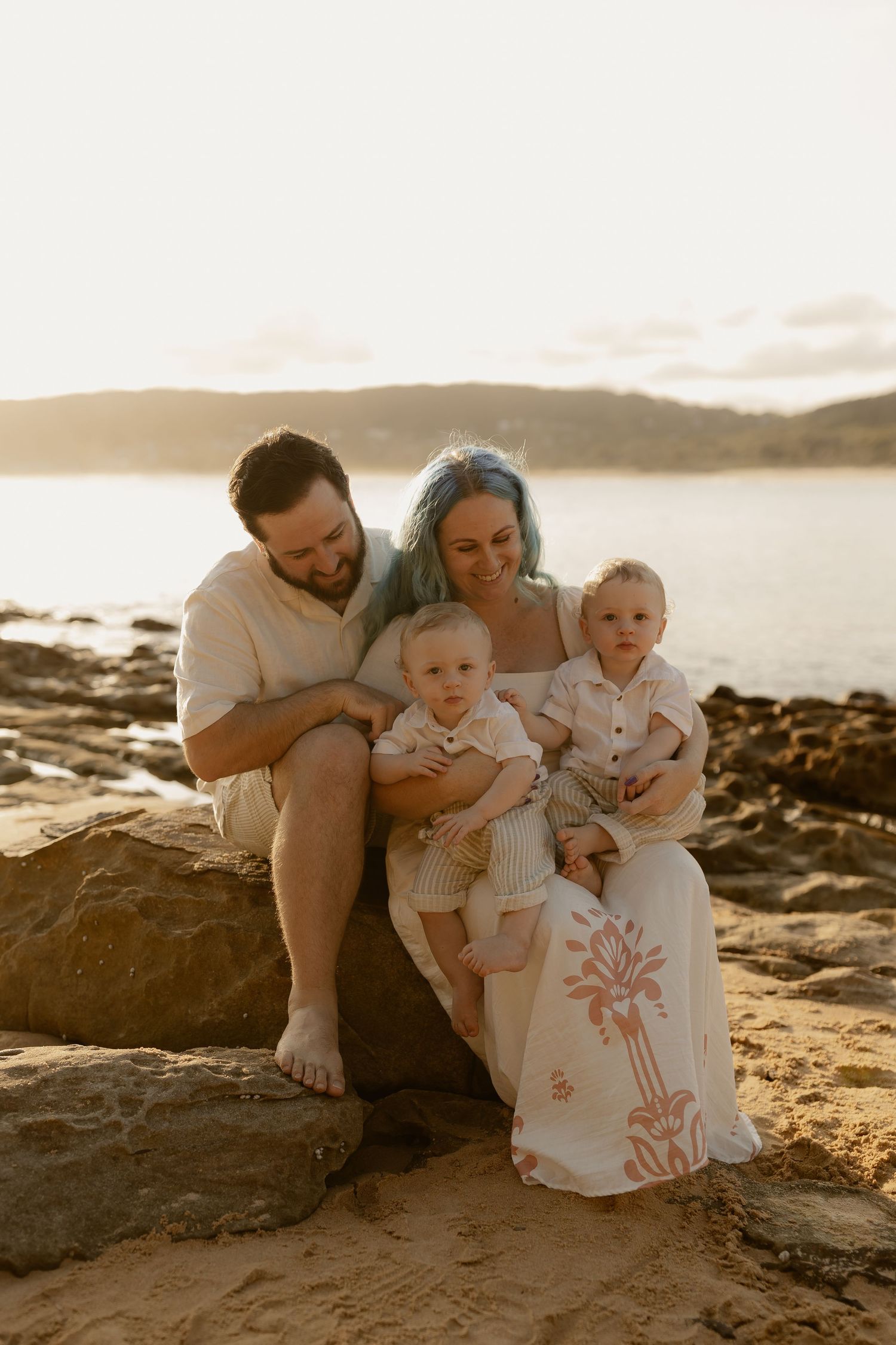 Family shares tender moments together on rocky beach at sunset with ocean views in background.