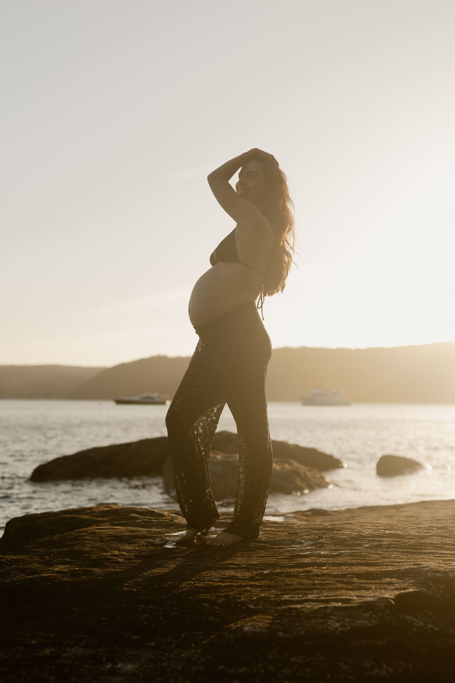A silhouetted figure sits on rocky coastal cliffs at sunset overlooking a tranquil ocean landscape.
