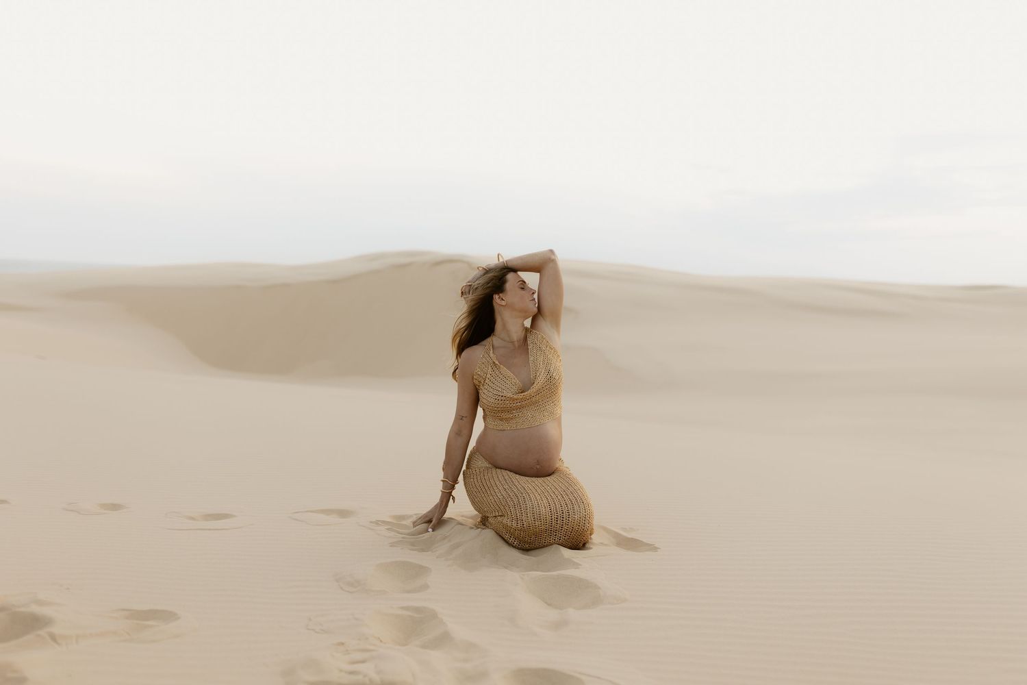 Figure in earth-toned attire sits thoughtfully among rolling desert sand dunes.