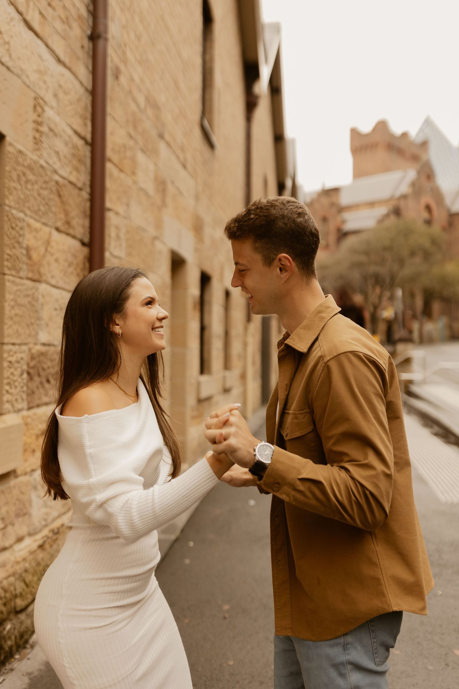 Couple holding hands and looking at each other while standing on a cobblestone street in front of historic buildings.