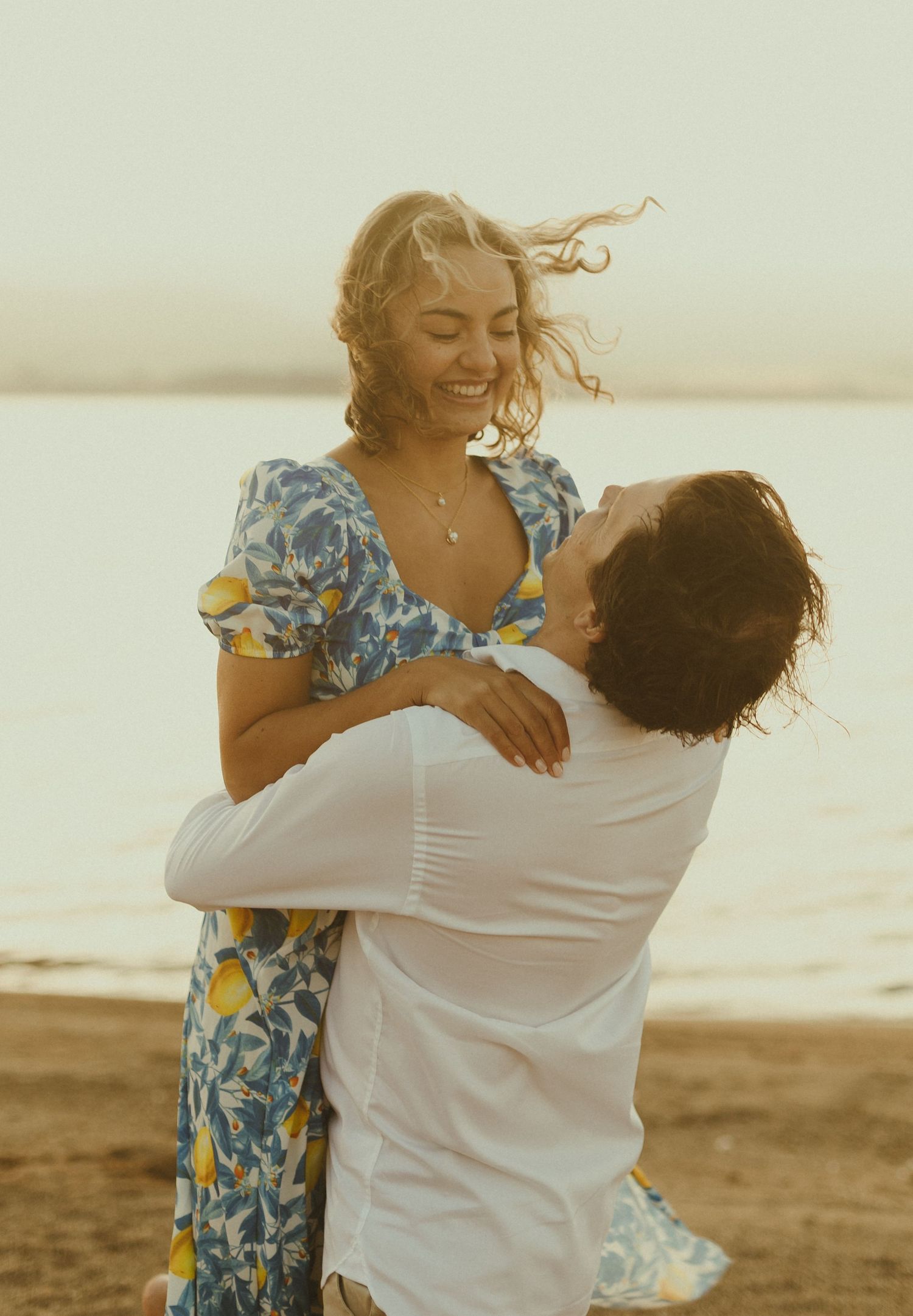 A couple embraces on a beach at sunset while the woman's floral dress and hair flow in the wind.