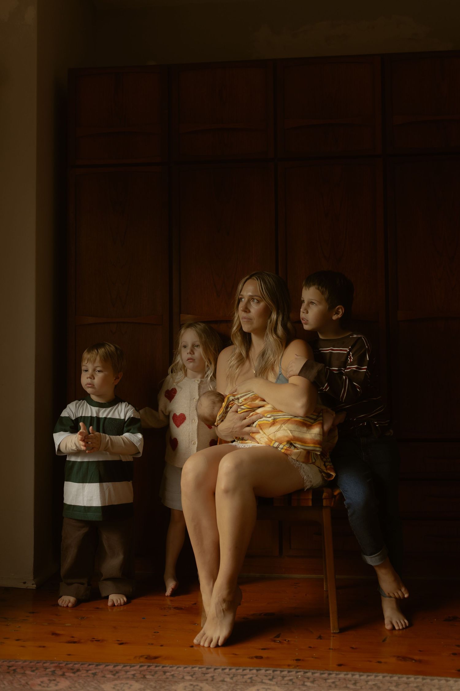 A mother sits on a chair breastfeeding her newborn, surrounded by her three young children in a warmly lit room.