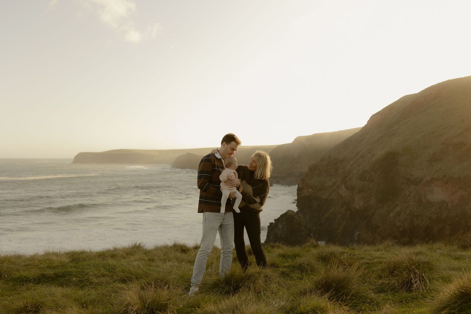 Young family with baby enjoying a scenic coastal clifftop view at golden hour sunset.
