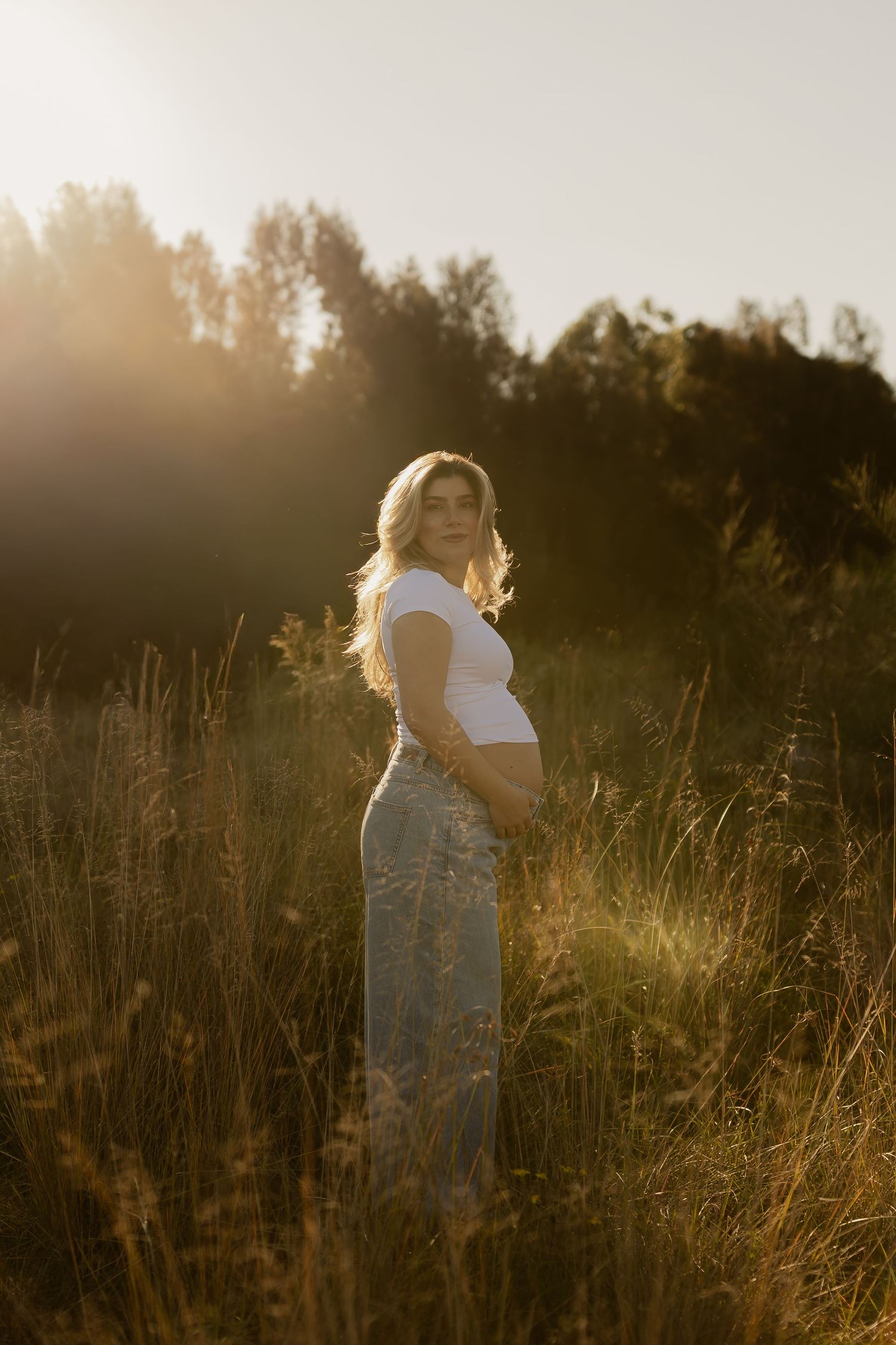 Pregnant woman in white crop top and blue skirt standing in golden field at sunset.