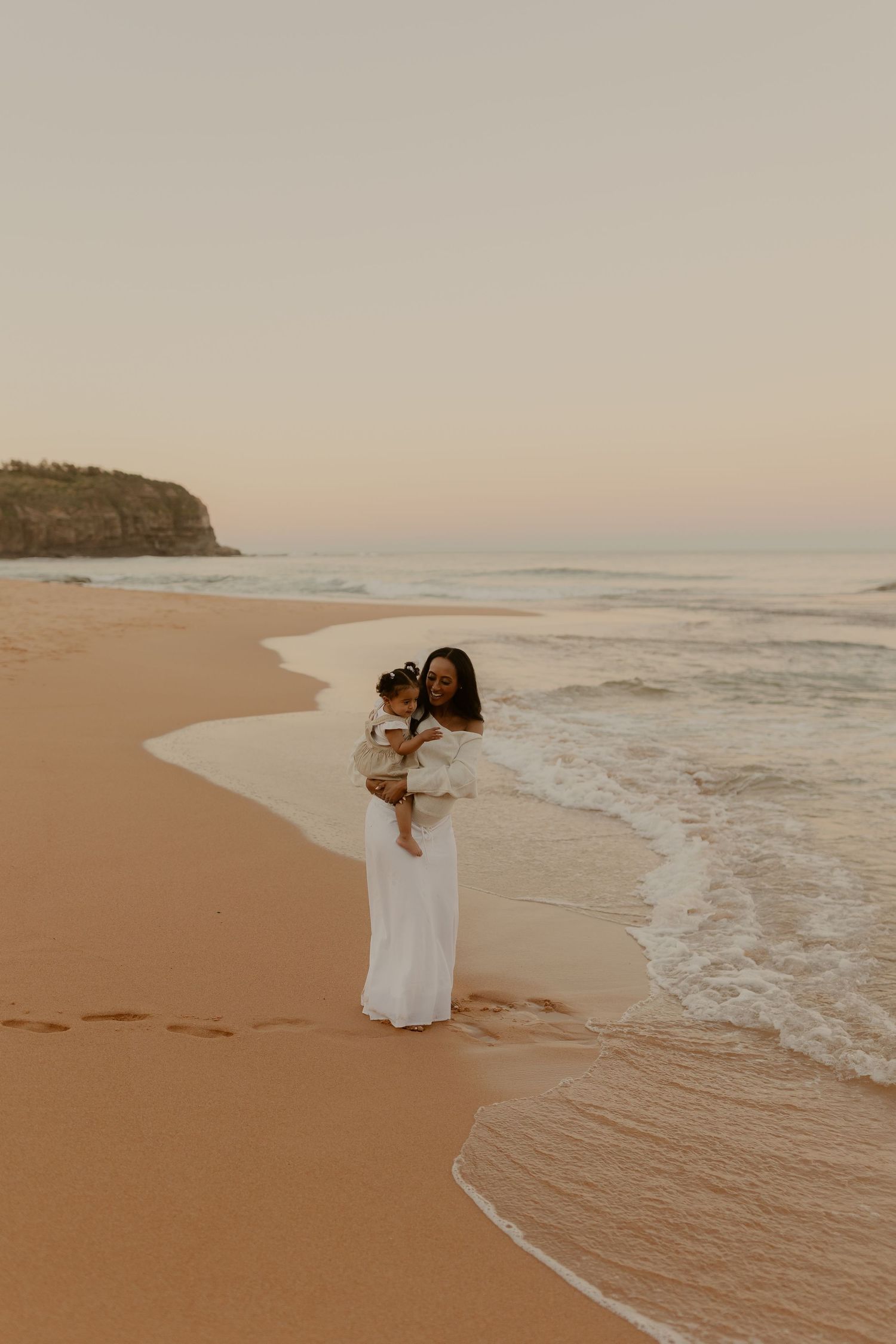 Mother in white dress holding baby on sandy beach at golden hour sunset by ocean waves.