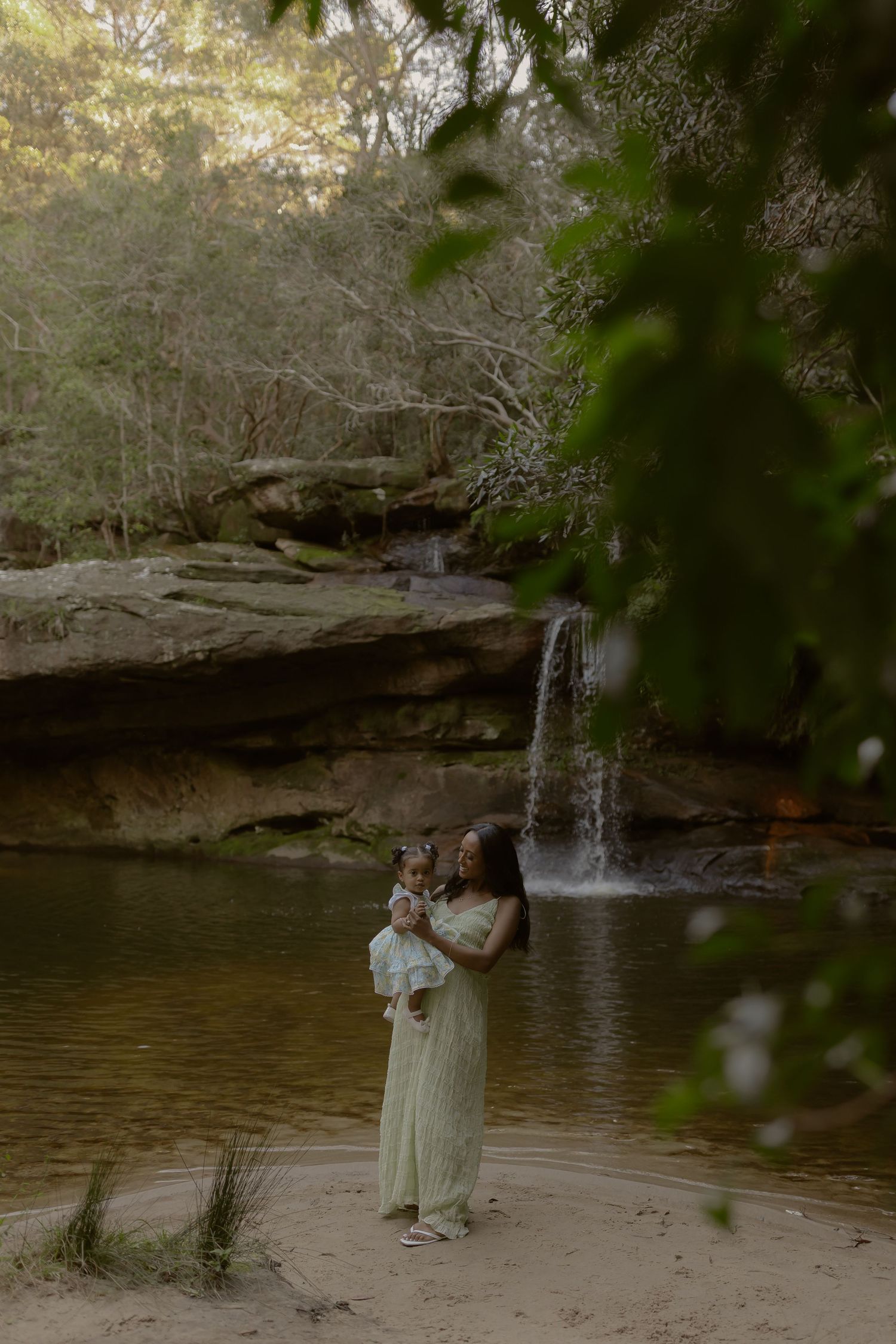 Woman in green dress holds baby near waterfall and rocky pool in lush forest setting.