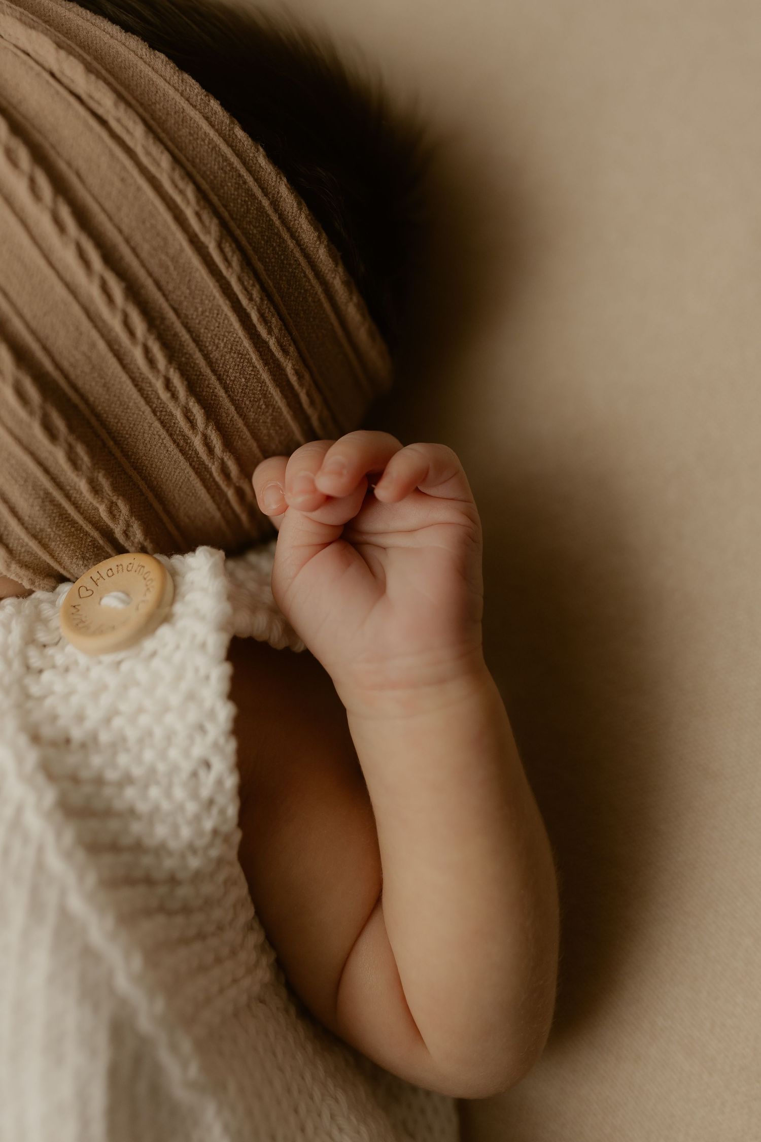 Close-up of a sleeping newborn baby's tiny fist in a cream knit romper with wooden button detail.