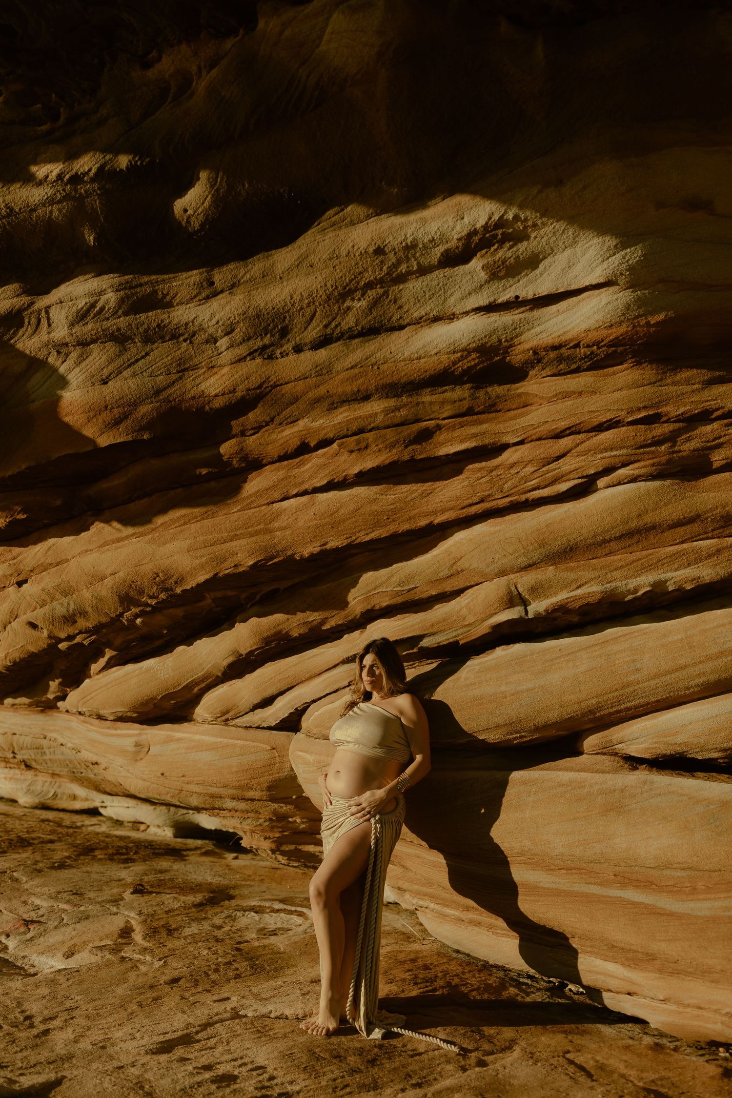 Pregnant woman in beige bikini posing against layered sandstone rock formations in a desert canyon setting.