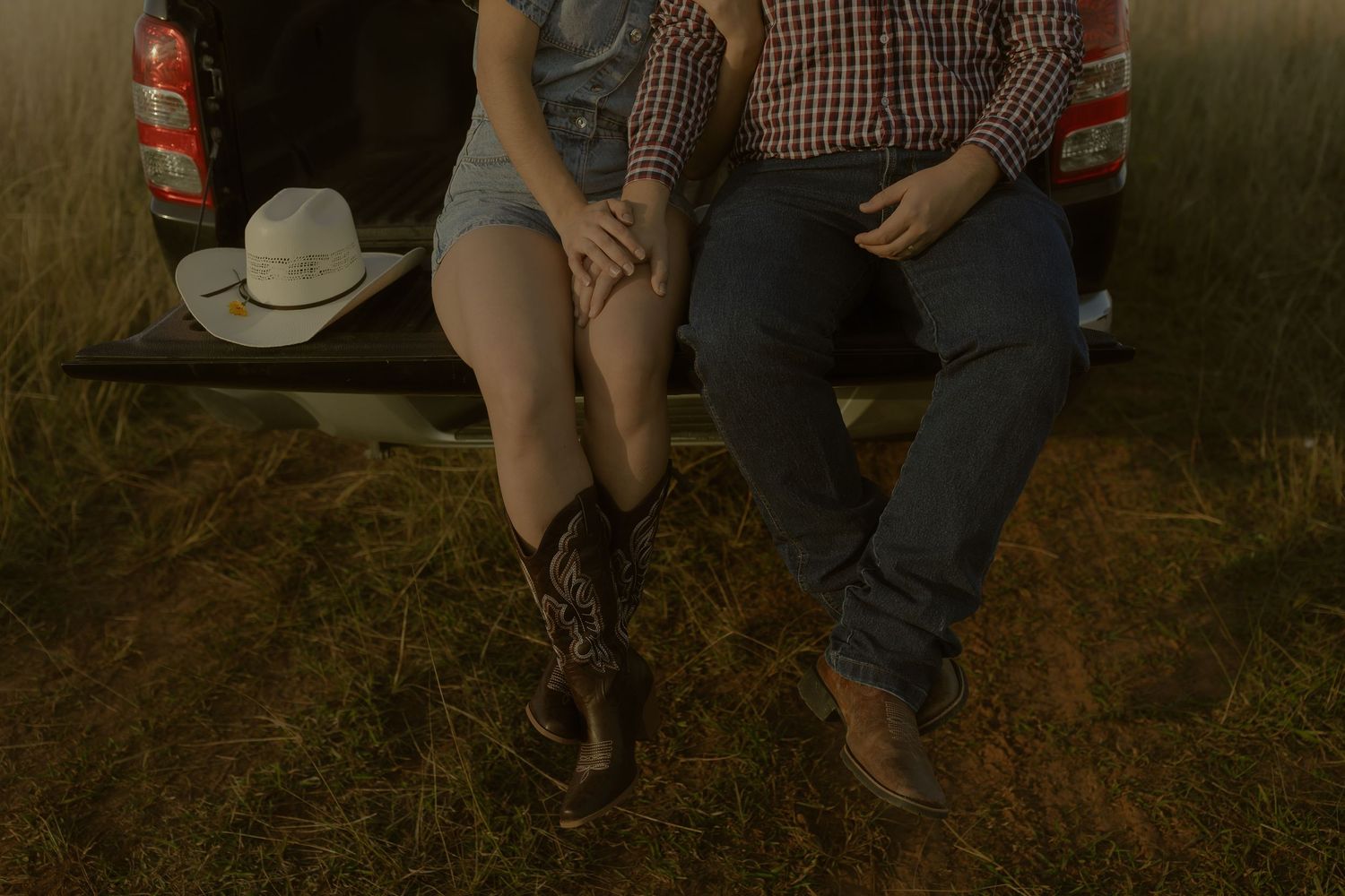 Couple sitting on a truck tailgate in a field, wearing cowboy boots and holding hands at sunset.
