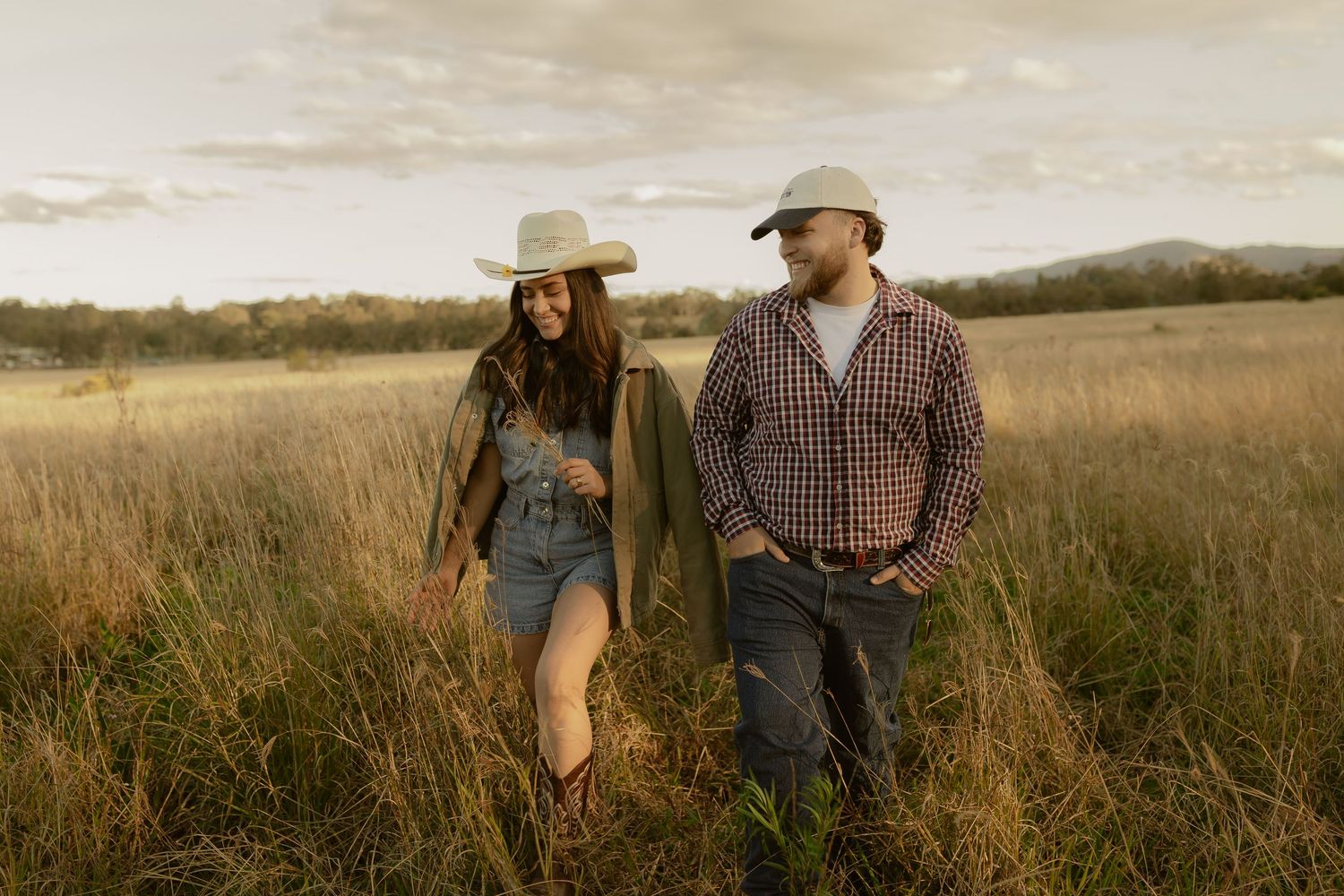 Couple in cowboy hats walking through golden field at sunset, wearing casual western outfits.