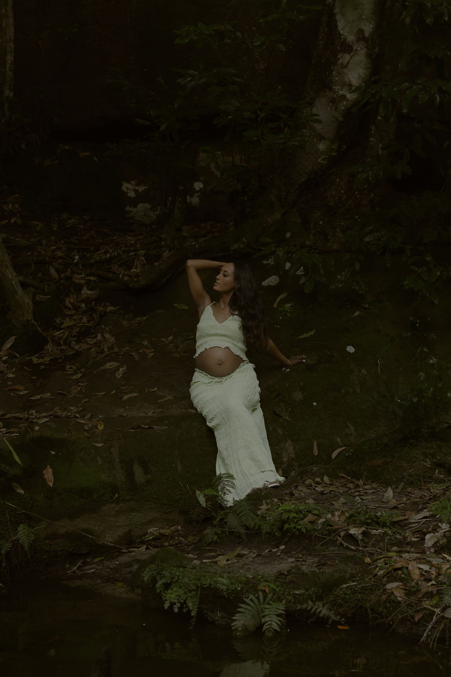 Pregnant woman in white dress poses gracefully in lush forest setting near water, head tilted back.