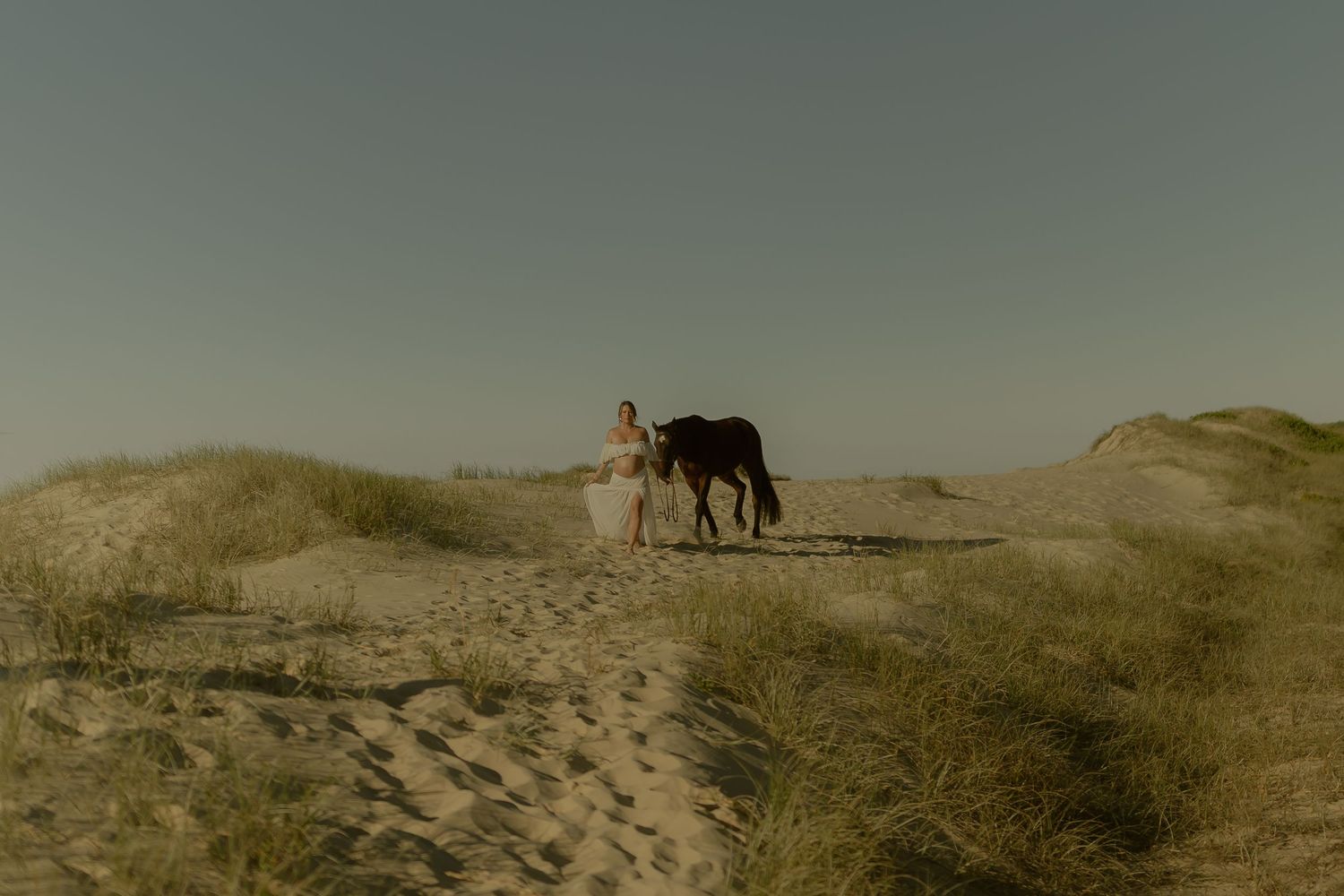 Woman in white dress walking with a dark horse on sandy dunes under a clear sky.