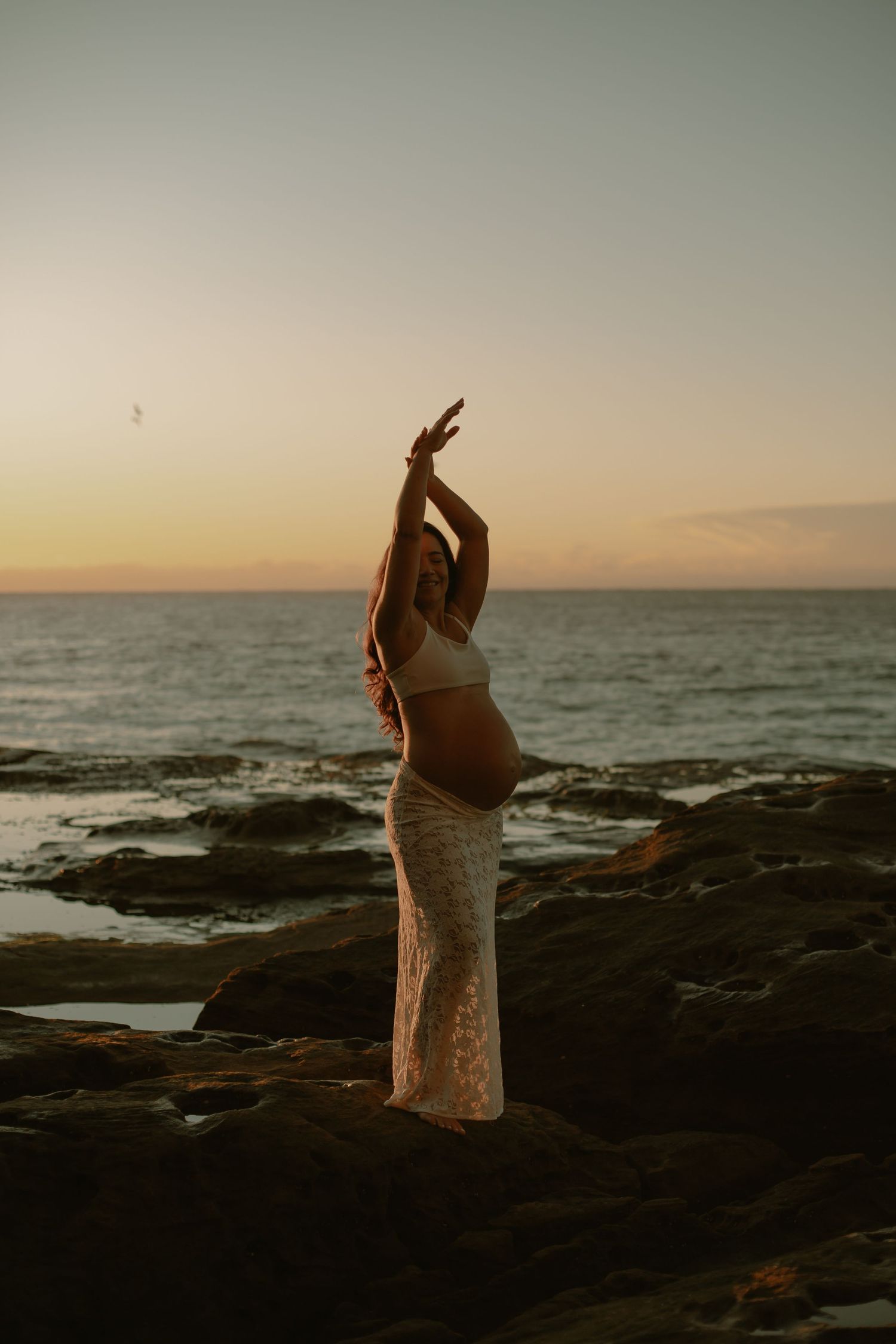 A figure in flowing lace dress poses gracefully against a sunset ocean backdrop on rocky shores.