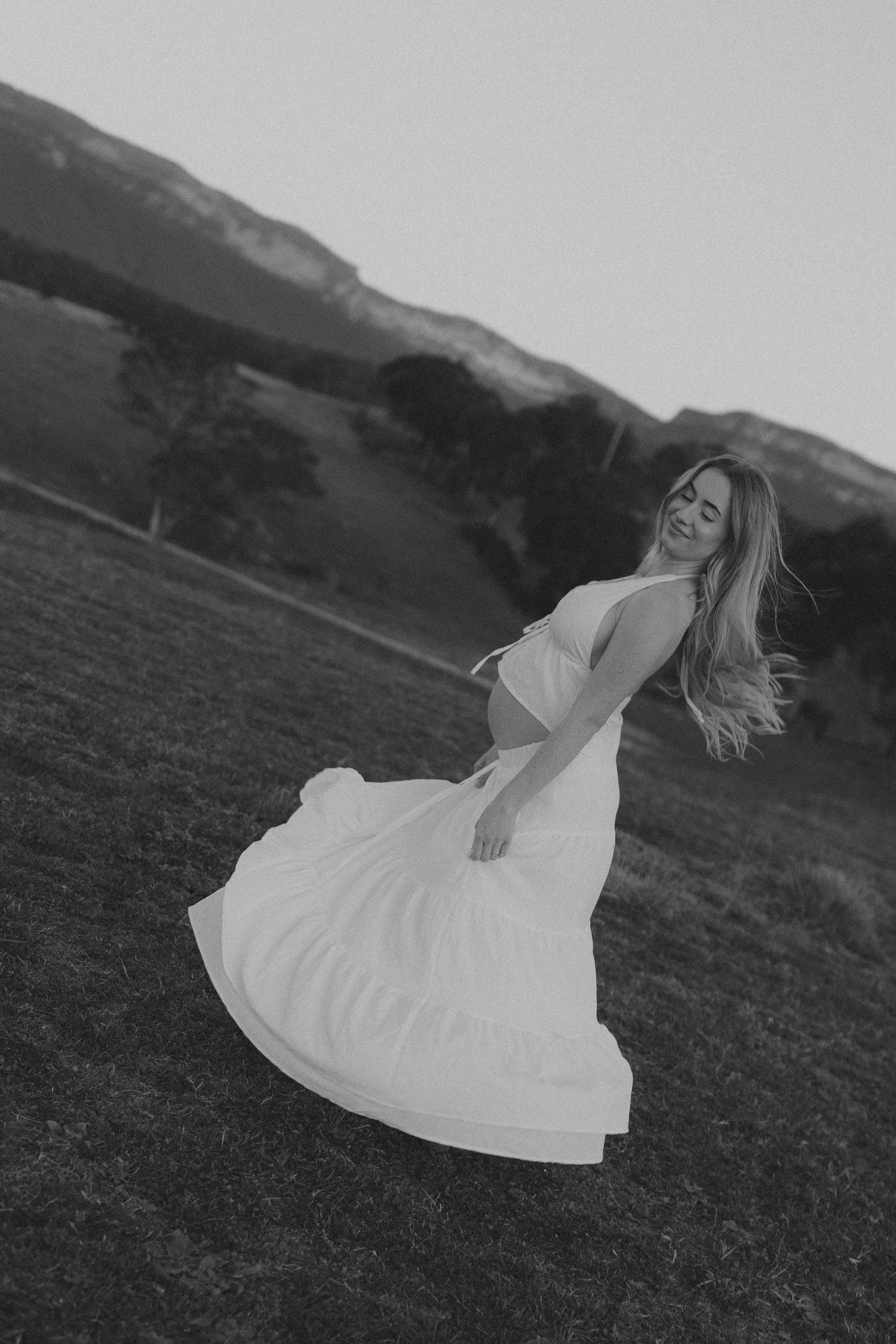 Black and white portrait of pregnant woman in white dress spinning in mountain meadow.