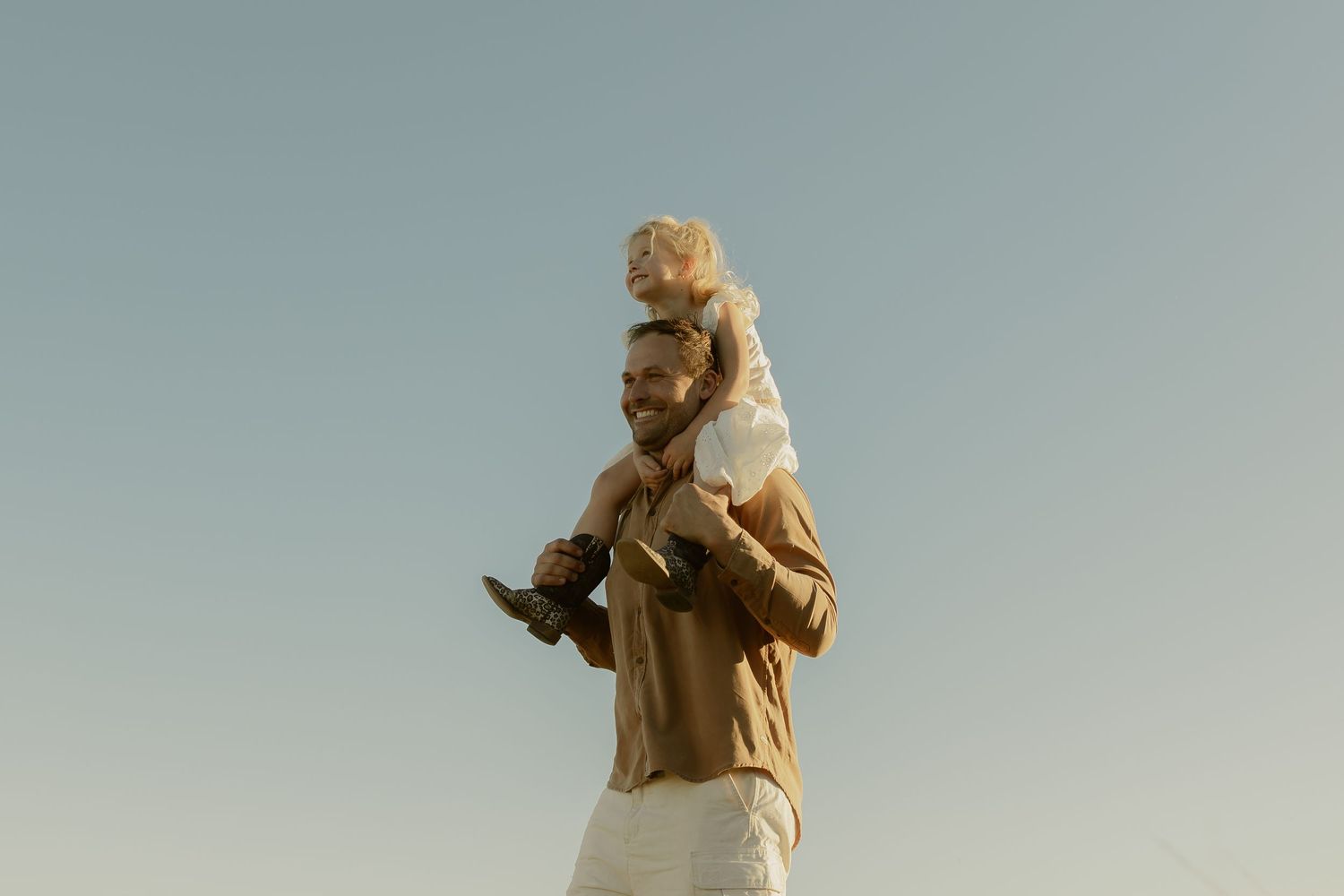 A man carries a young child on his shoulders against a clear blue sky, both enjoying a sunny outdoor moment.