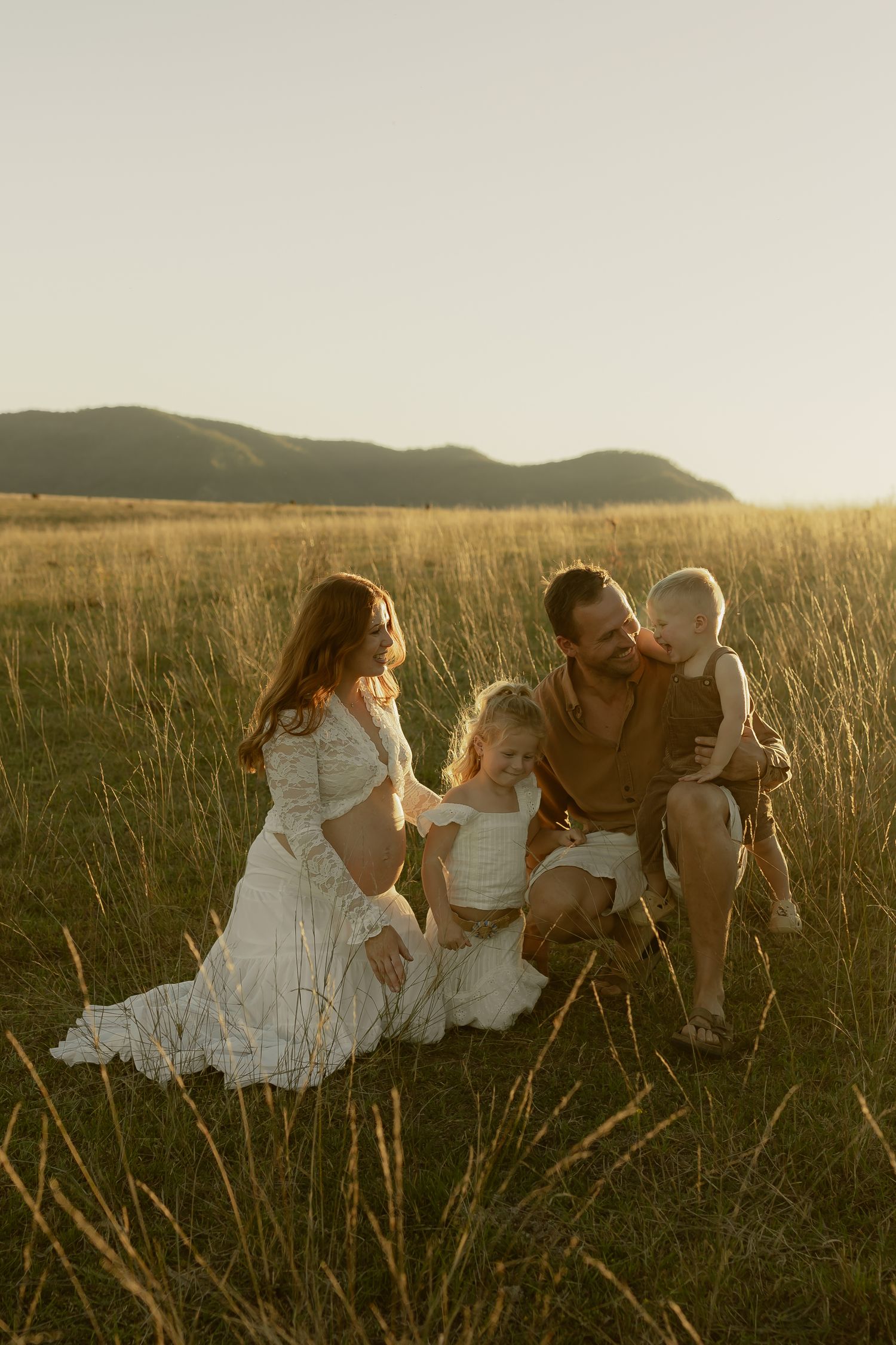 Family of four enjoying golden hour in a grassy field with mountain backdrop, mother in white dress, father holding toddler.