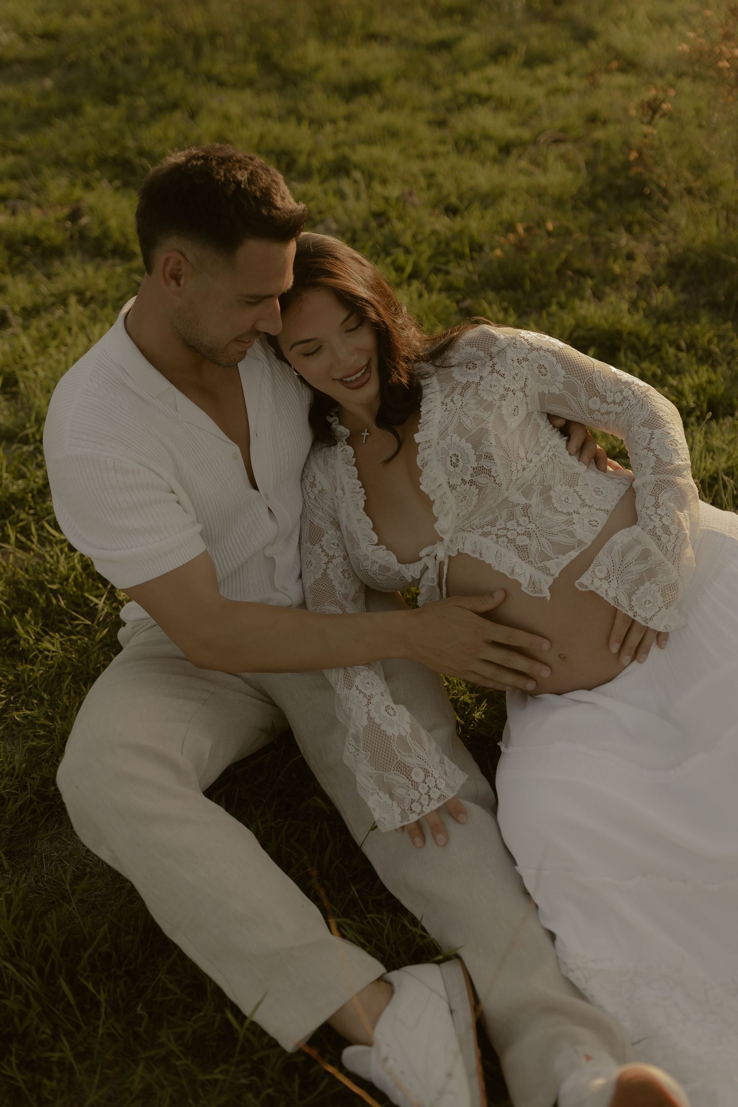 A tender moment between a couple lying in a sunlit meadow wearing white attire.