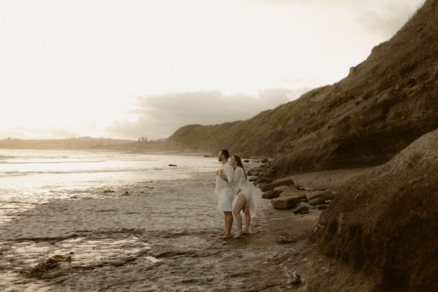 Serene beach landscape with silhouettes against setting sun and rocky coastal formations in muted tones.
