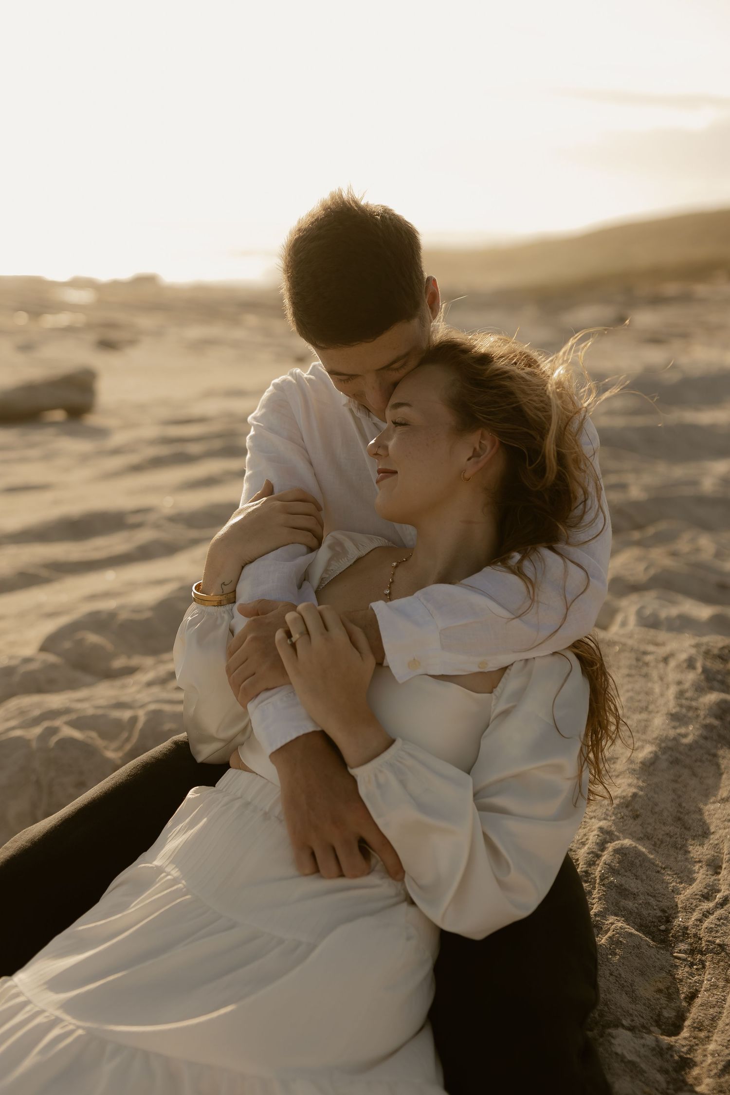 Intimate couple embracing on sandy beach during golden hour in white wedding attire.