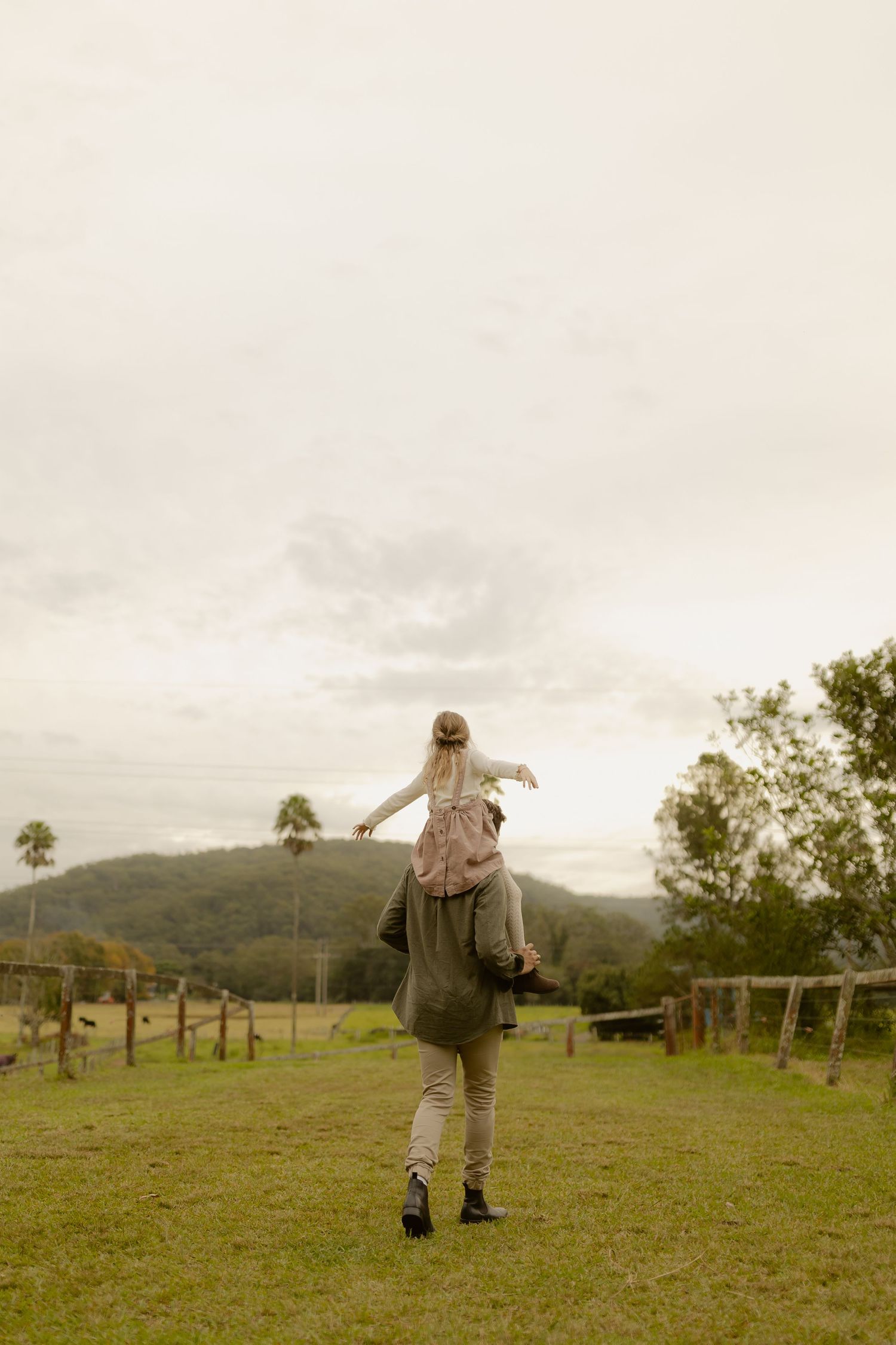 Child with arms outstretched rides on adults shoulders walking through a scenic green farm landscape.