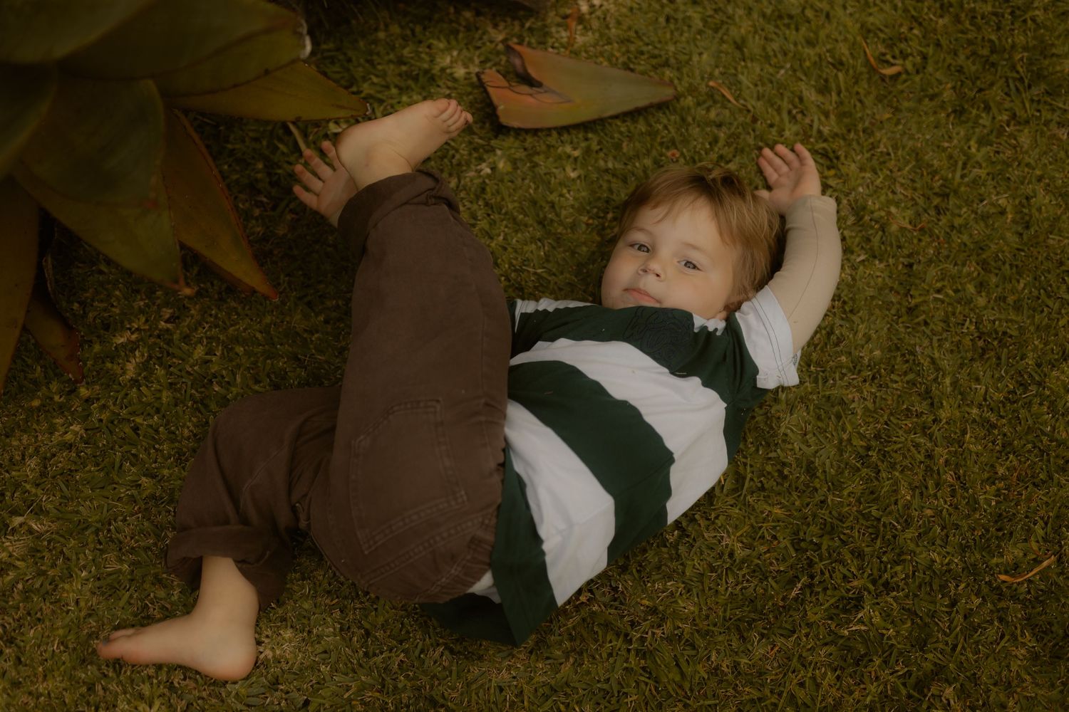 Young toddler boy lying on green grass outdoors wearing striped shirt and brown pants.