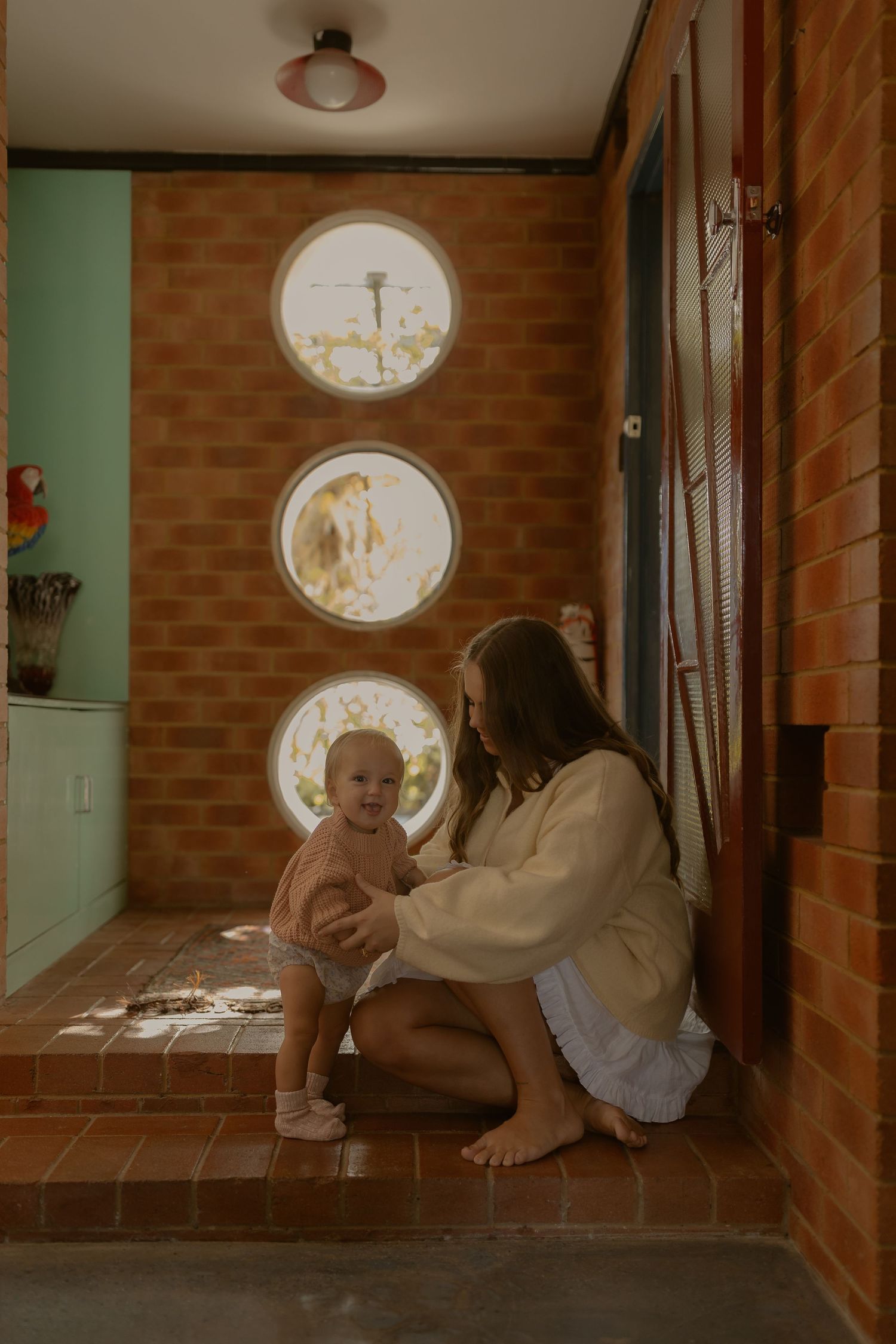Mother sitting on brick floor playing with smiling baby near round porthole windows in a warm terracotta interior.