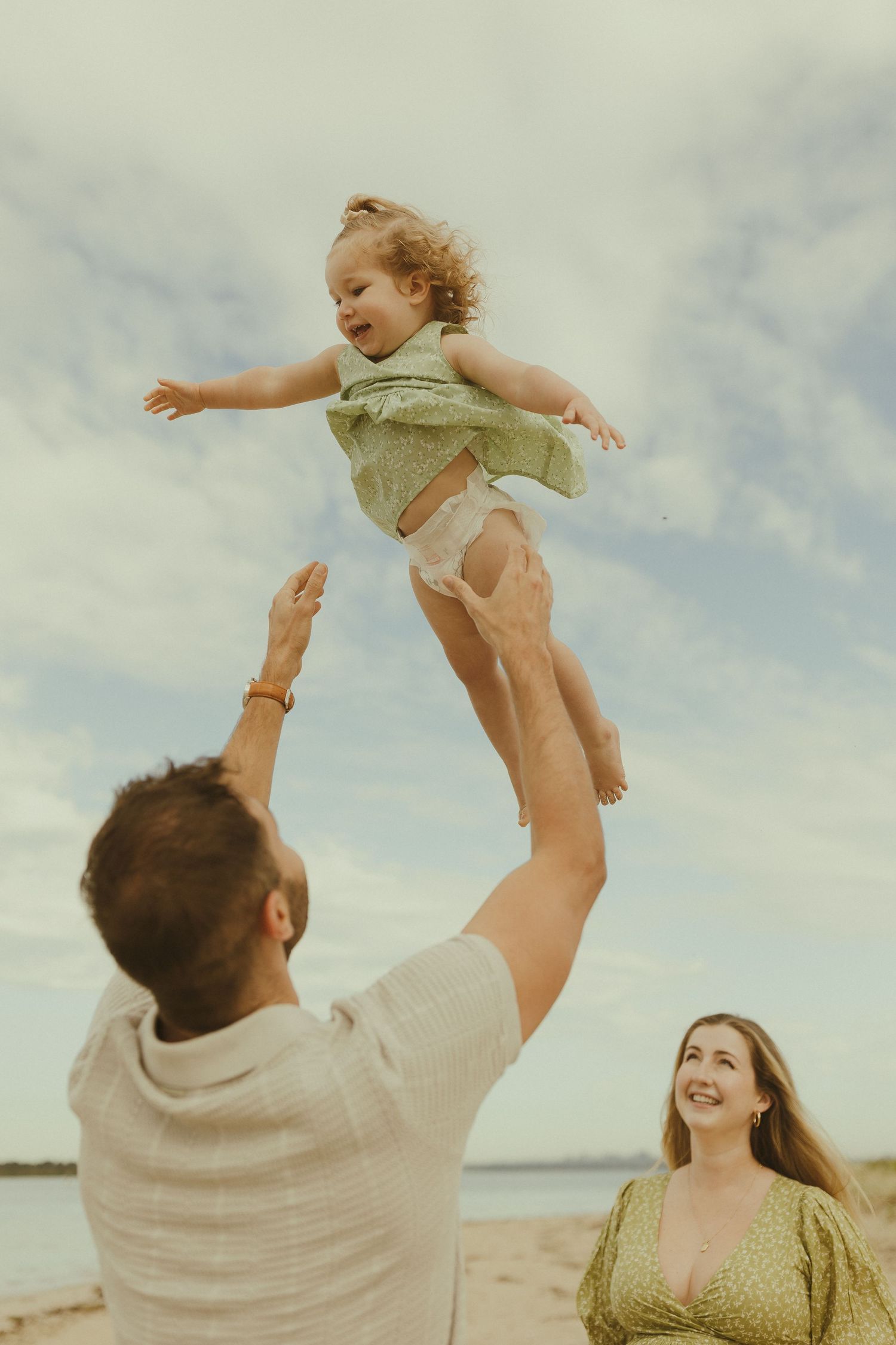 A family plays together on a sunny beach, lifting a small child into the air with joyful expressions.