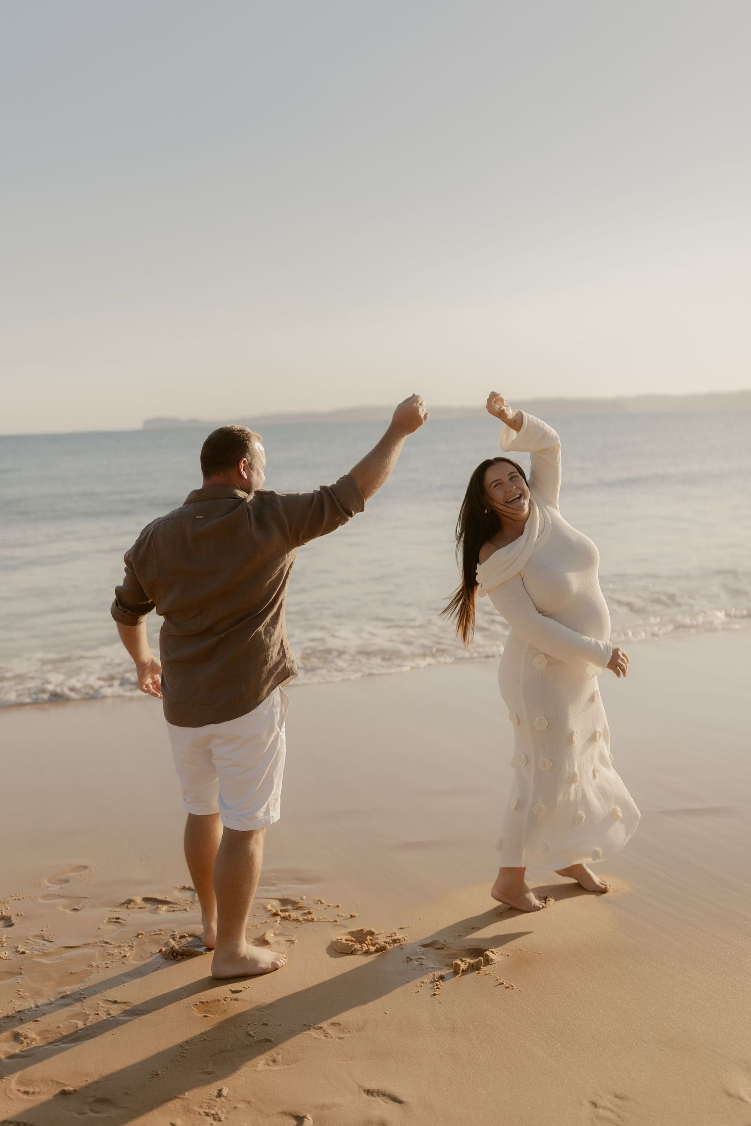 Two people dance joyfully on a sunlit beach at golden hour.