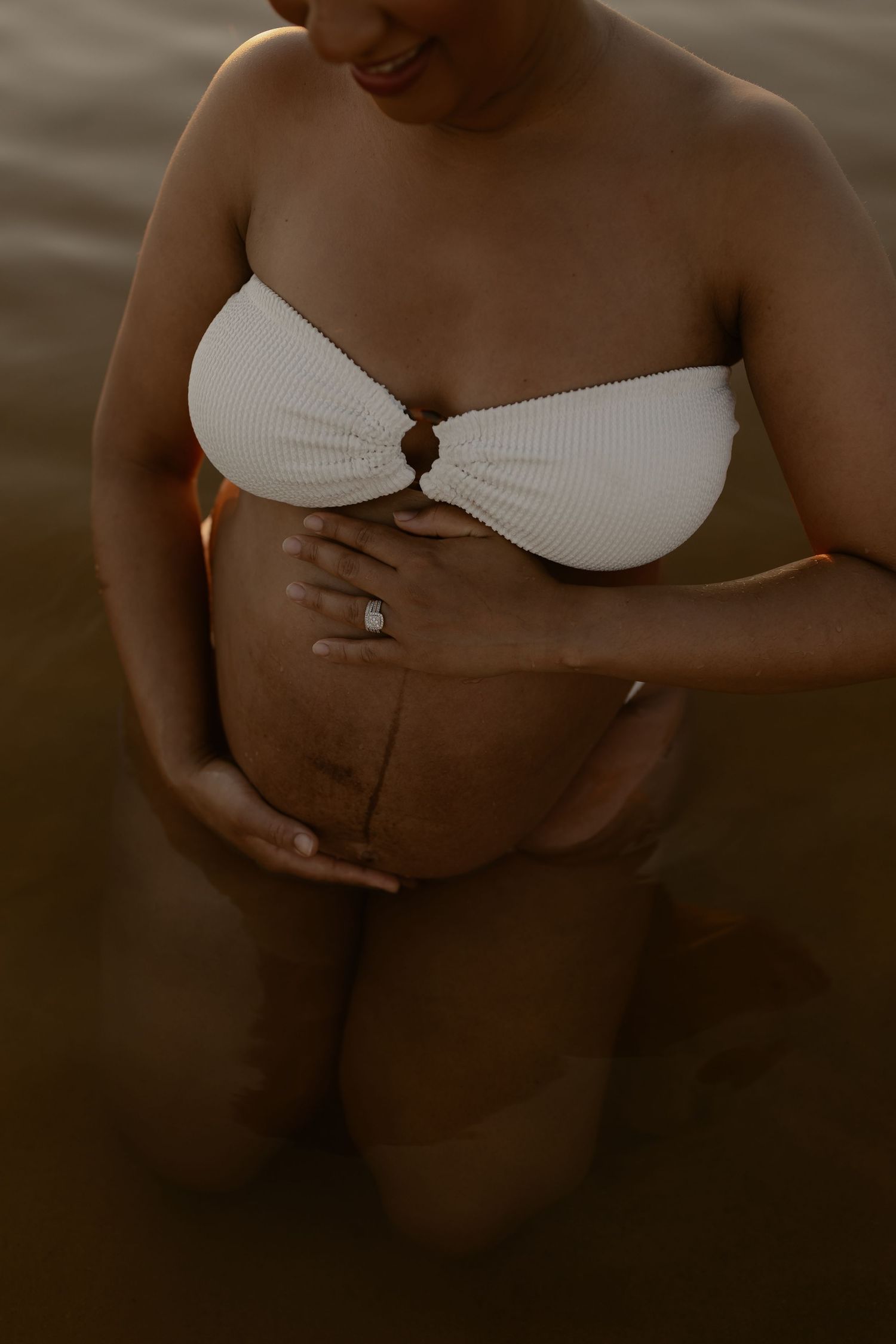 Artistic maternity photo showing pregnant woman in white bikini top standing in water during golden hour.