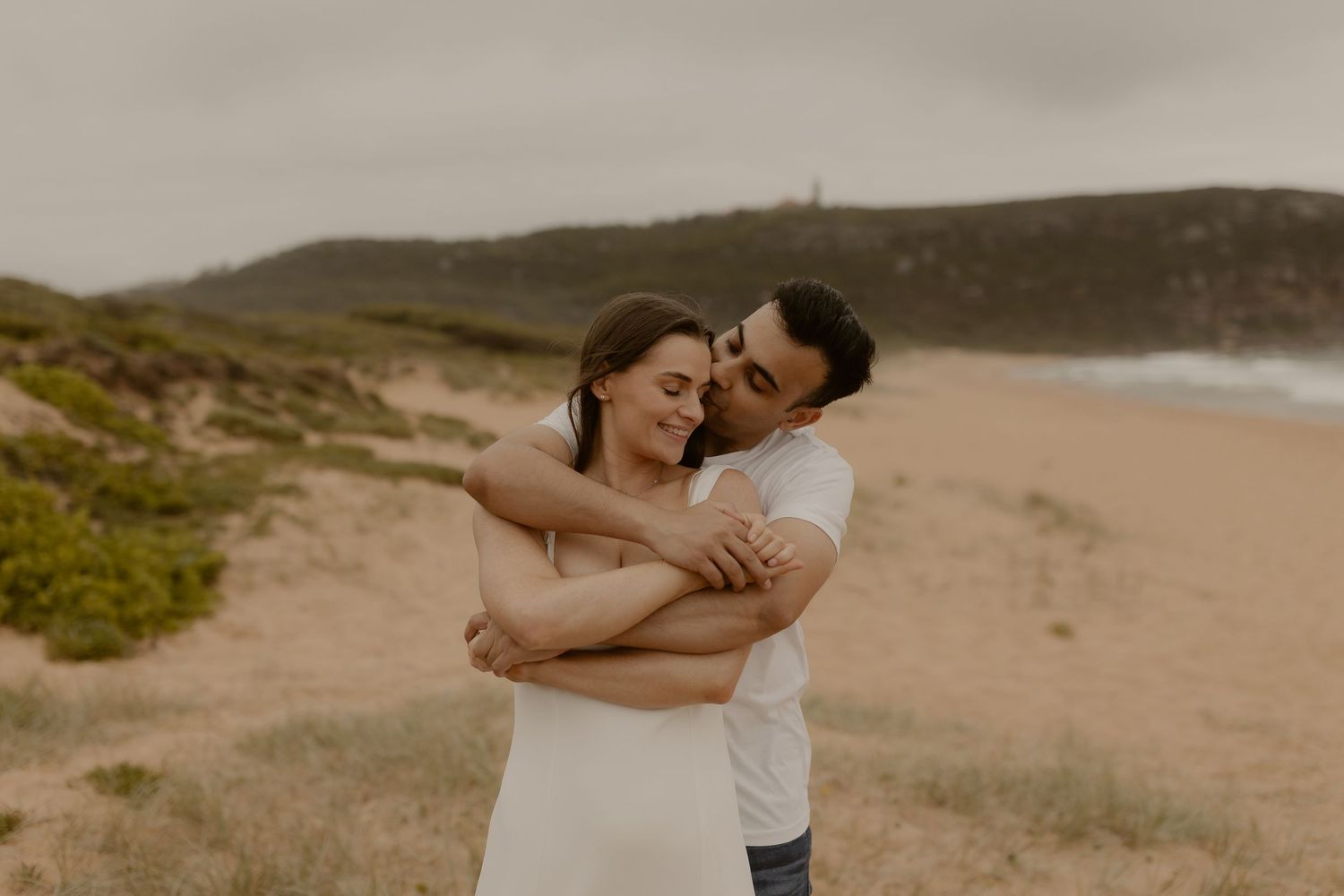 Two people embrace lovingly on a scenic beach with coastal cliffs in the background.
