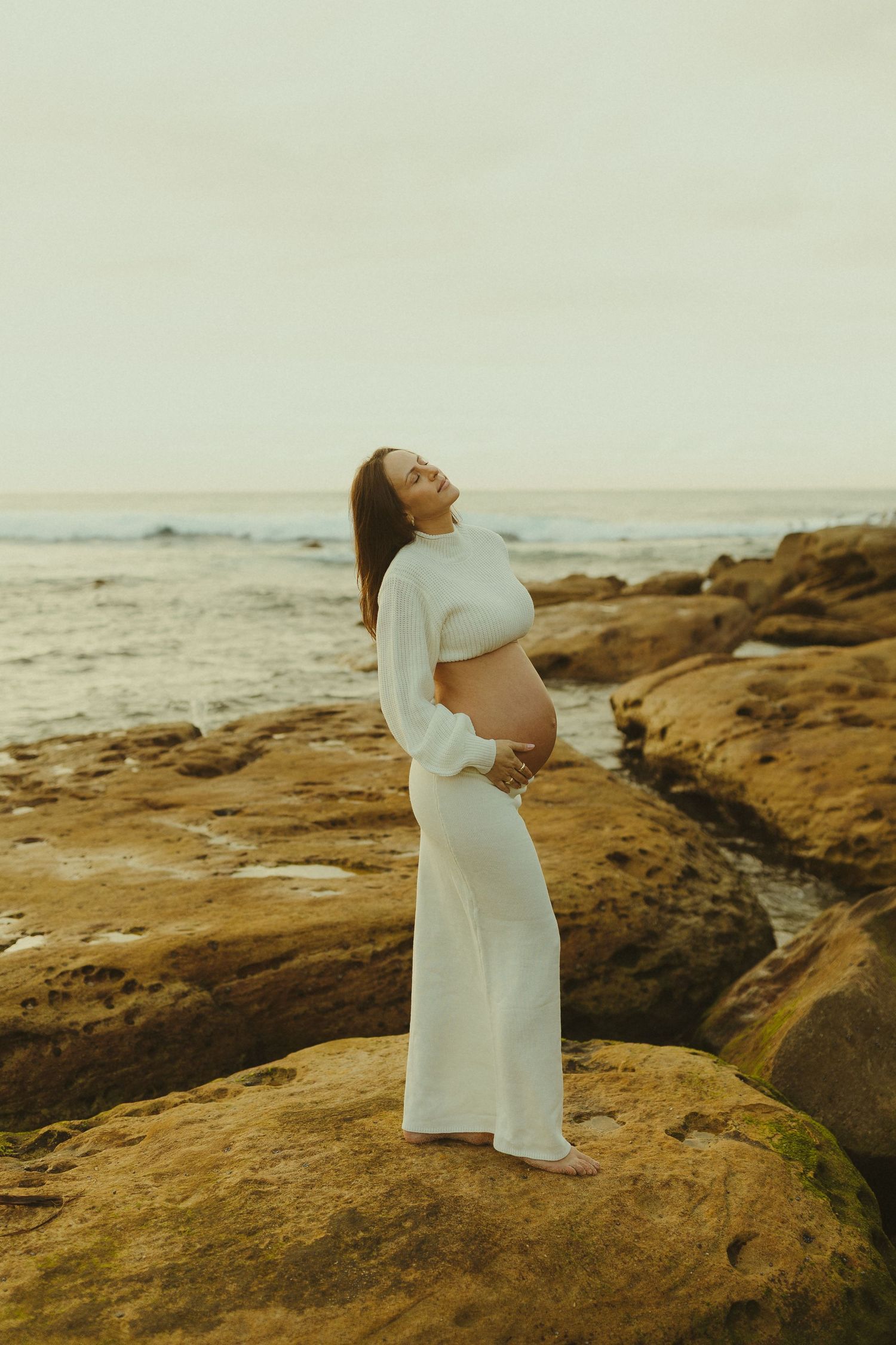 A person in white stands gracefully on coastal rocks with ocean waves in the background.