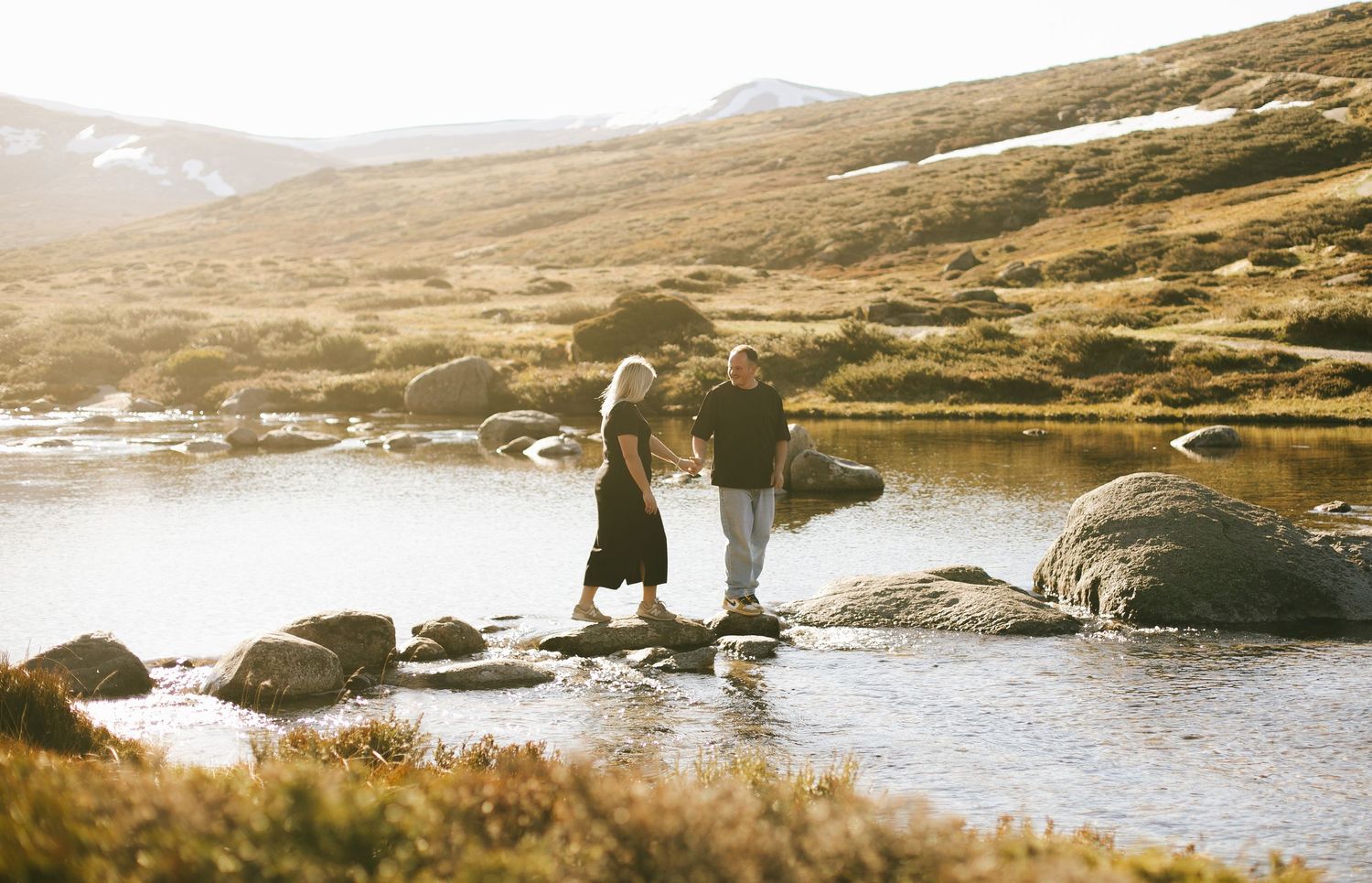 Two figures stand together on rocks by a mountain lake at sunset in a serene landscape.