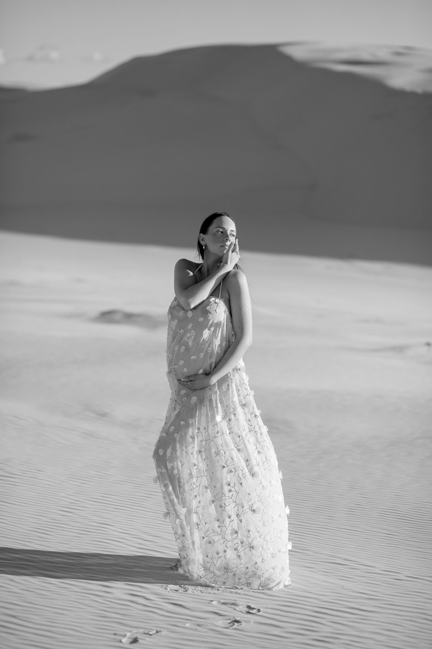 Person in white lace dress poses elegantly among rolling sand dunes under cloudy sky in black and white photo.