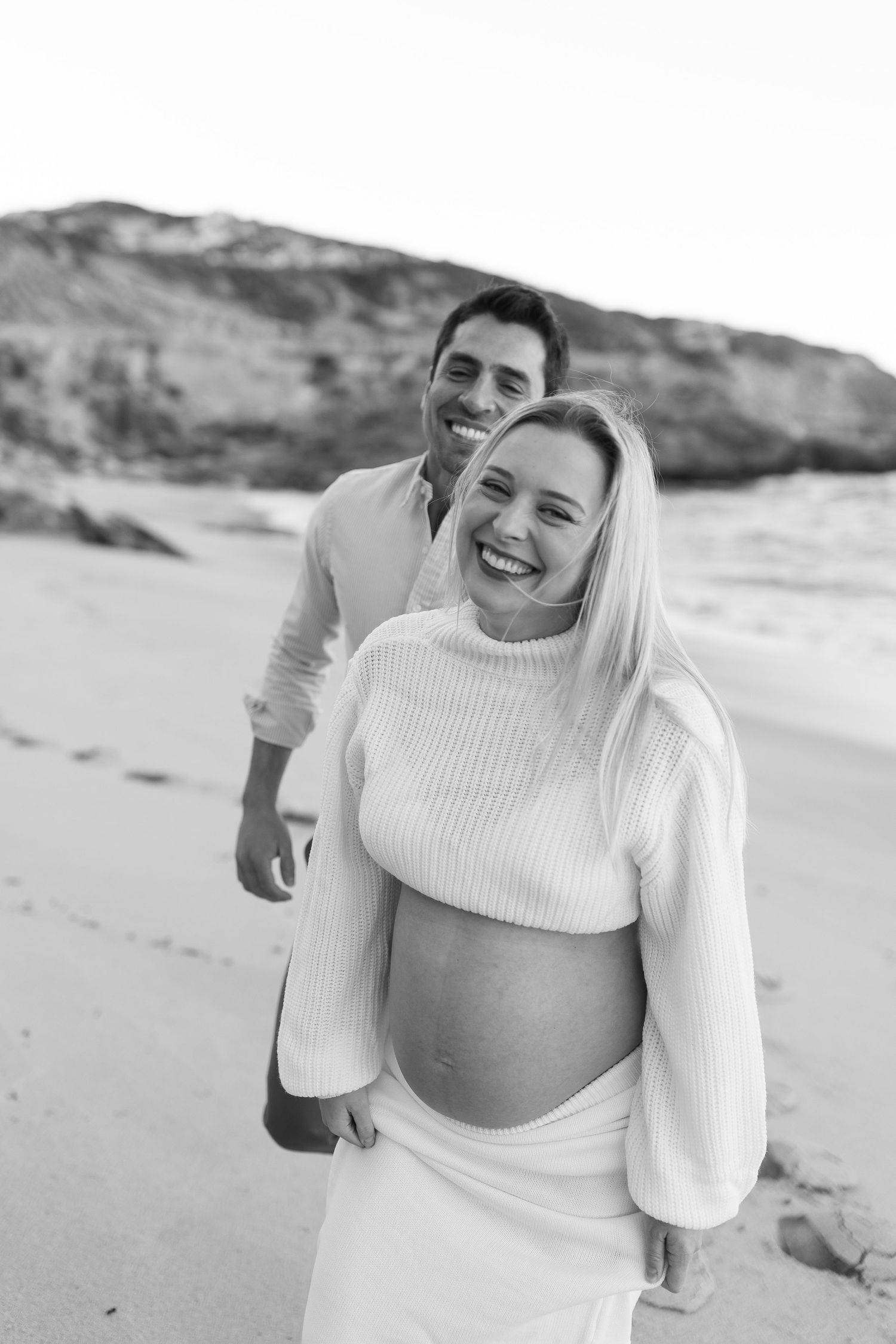 Black and white portrait of an expecting couple laughing together on a beach with coastal cliffs in background.